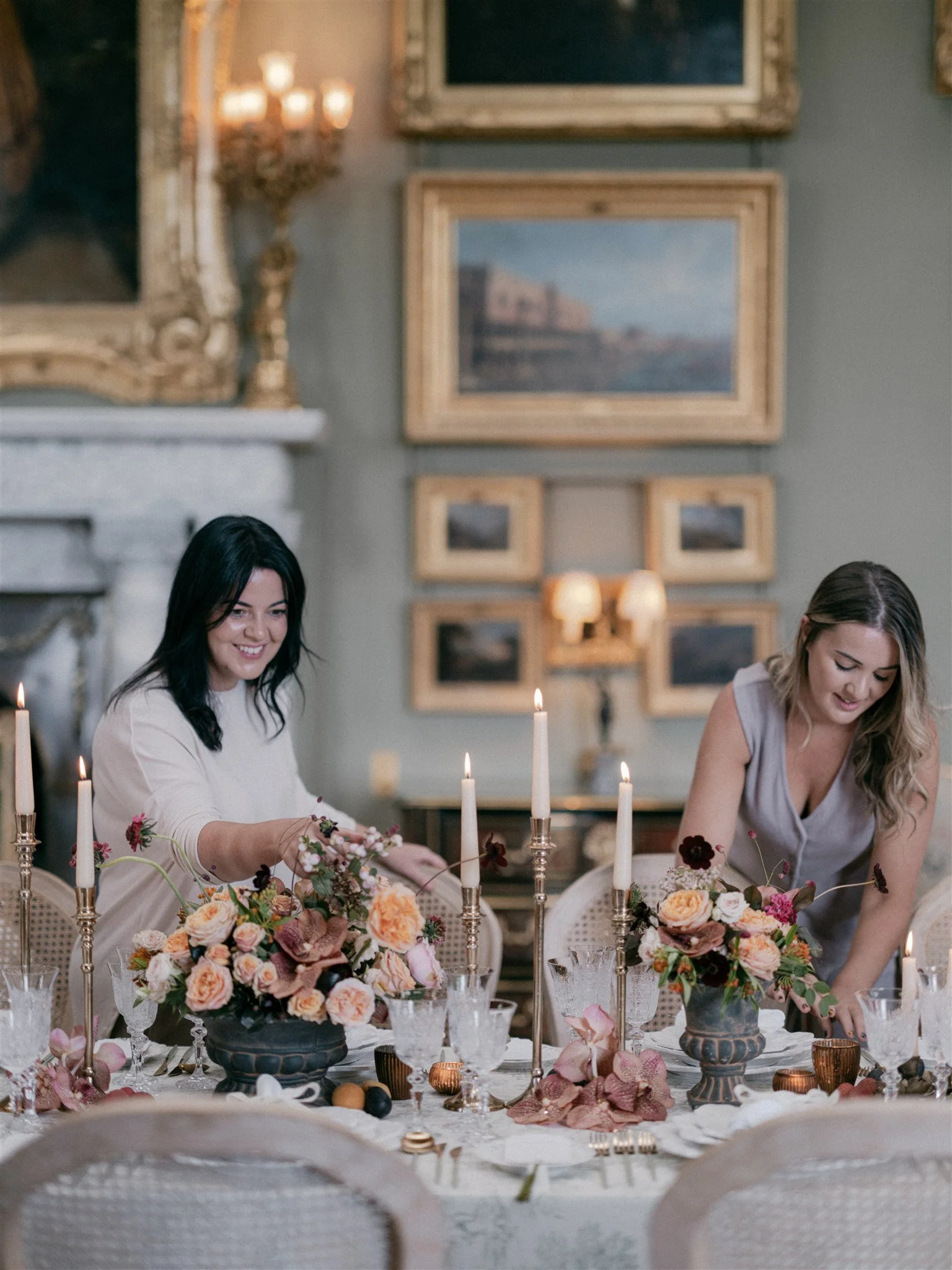 Two women setting a floral centerpiece on a decorated dining table in an elegant room.