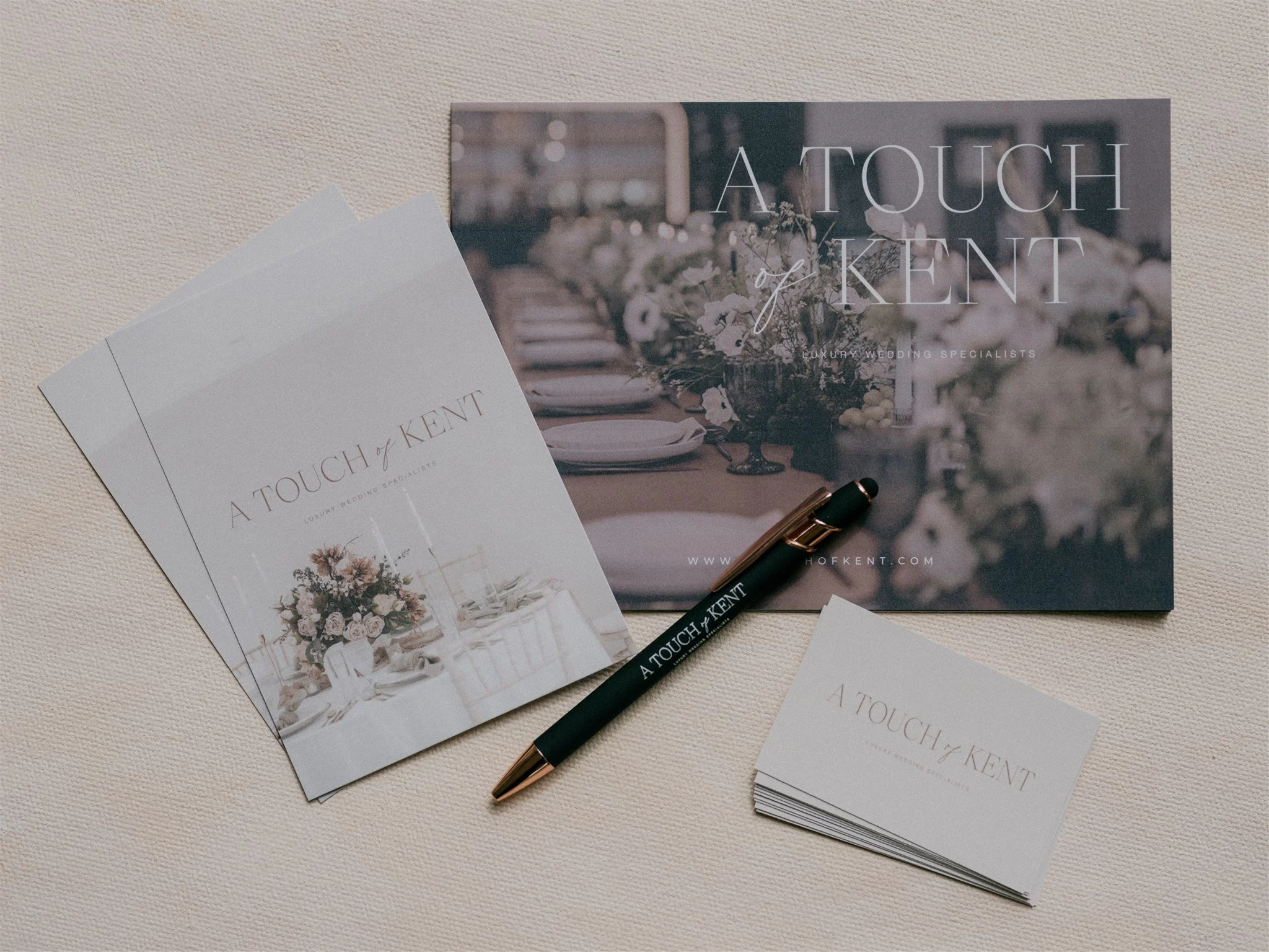 Wedding promotional materials including brochures, a pen, and business cards from A Touch of Kent, on a beige textured surface.
