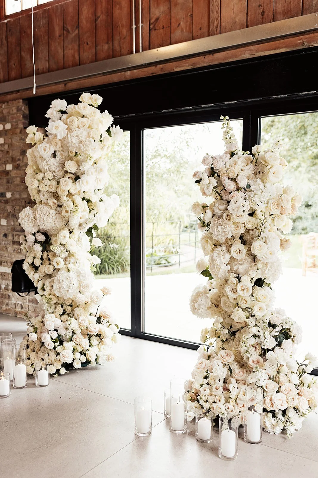 Two large floral arrangements made of white roses and other white flowers in front of a glass door, with candles in glass holders on the floor.