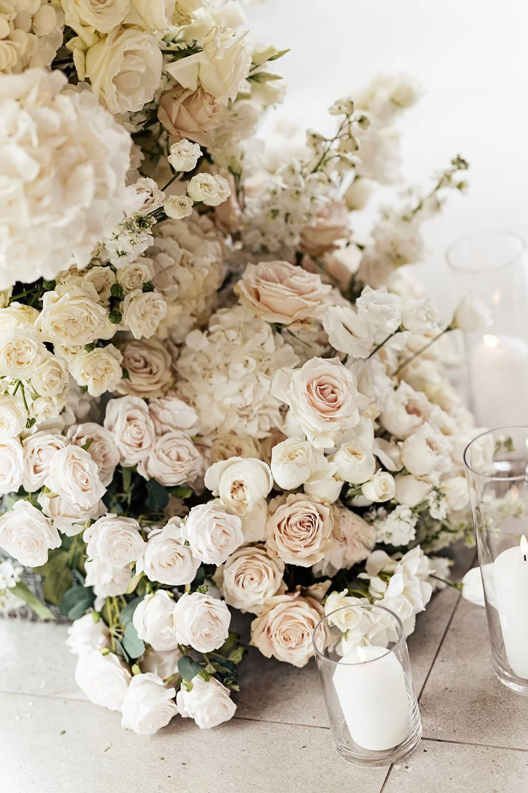 A large arrangement of white and pale pink roses and hydrangeas on a table, with white candles in glass holders.