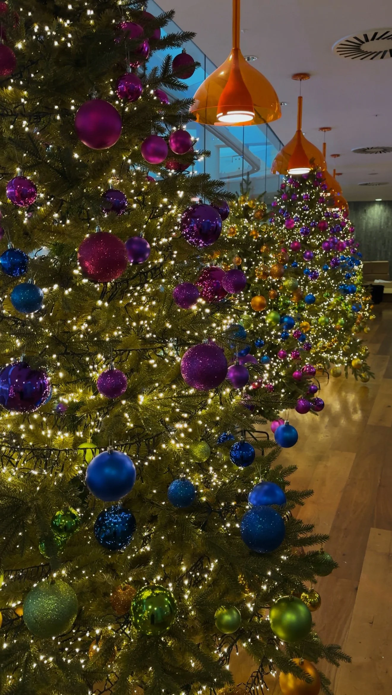 Decorated Christmas trees with baubles in purple, blue, green, and gold, illuminated by string lights, in a festive indoor setting.