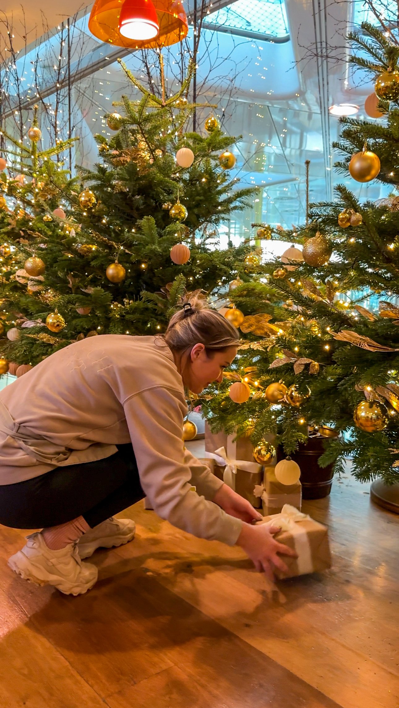 A woman is crouching down, placing a wrapped gift under a decorated Christmas tree with gold and white ornaments, surrounded by wrapped presents, inside a room with a glass ceiling and string lights.