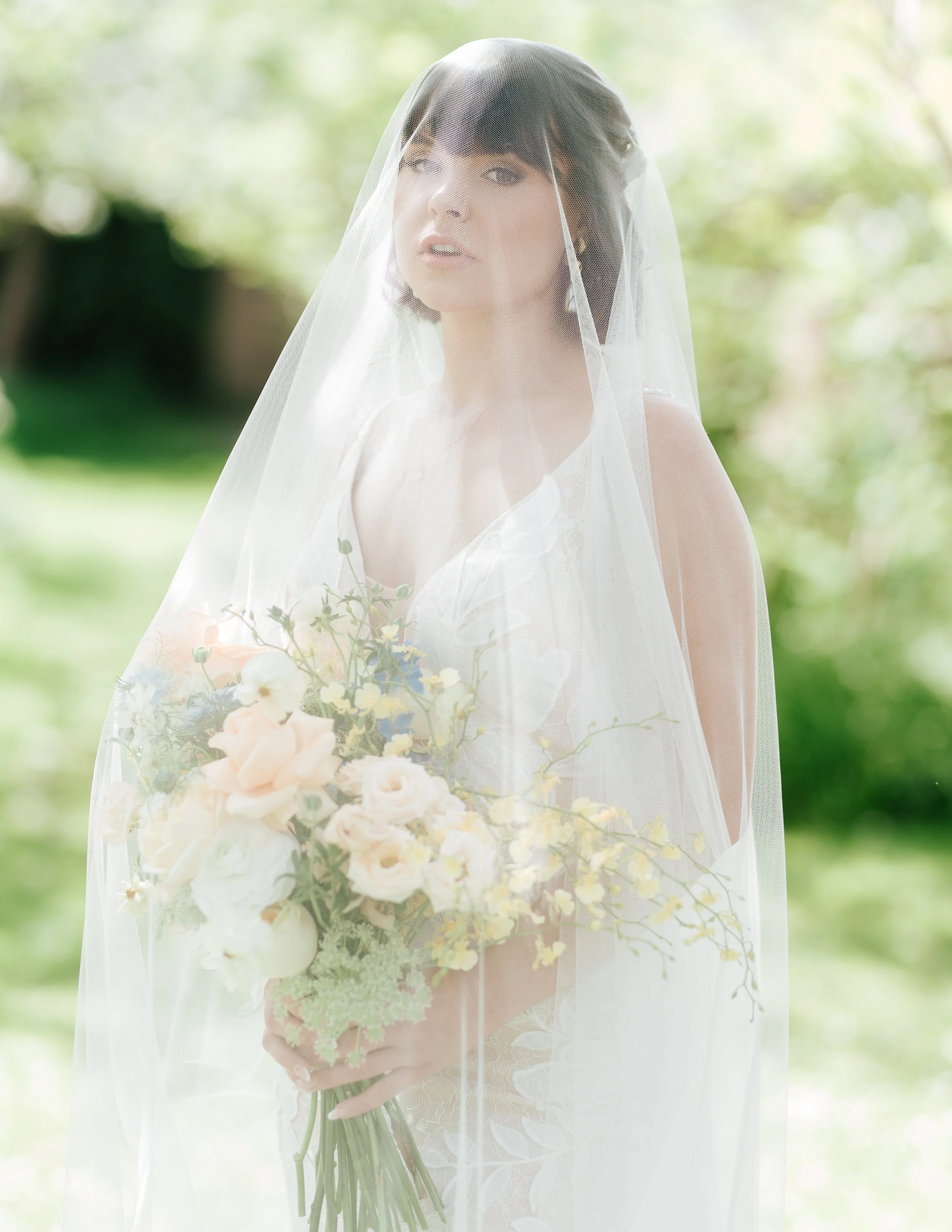 A bride wearing a lace wedding dress and a sheer veil, holding a bouquet of pale pink, white, and yellow flowers, with a soft outdoor background.