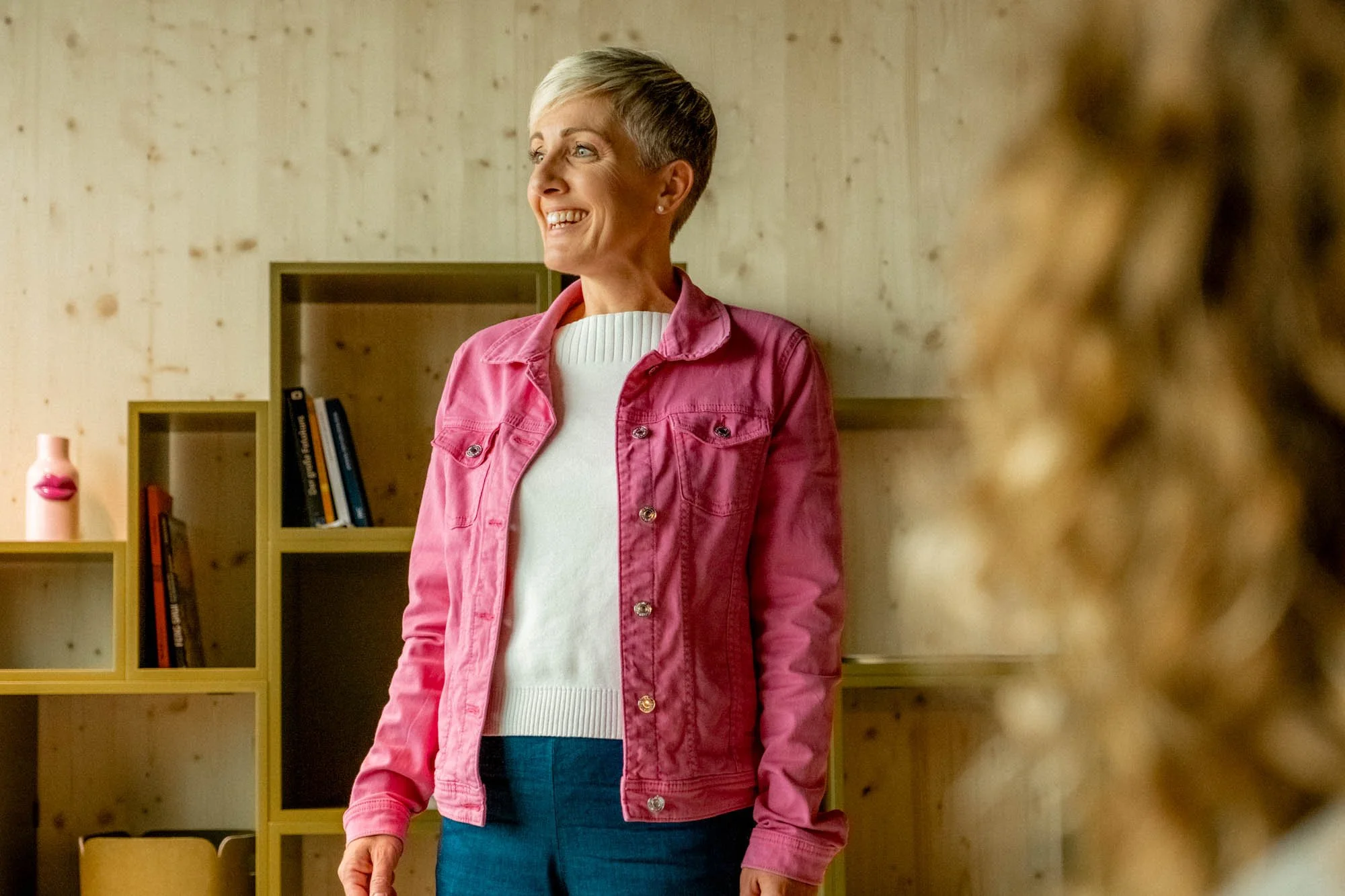 Gedächtnistrainerin Petra Oggenfuss in pinker Jacke am Lächeln. Steht vor einer Holzwand am präsentieren