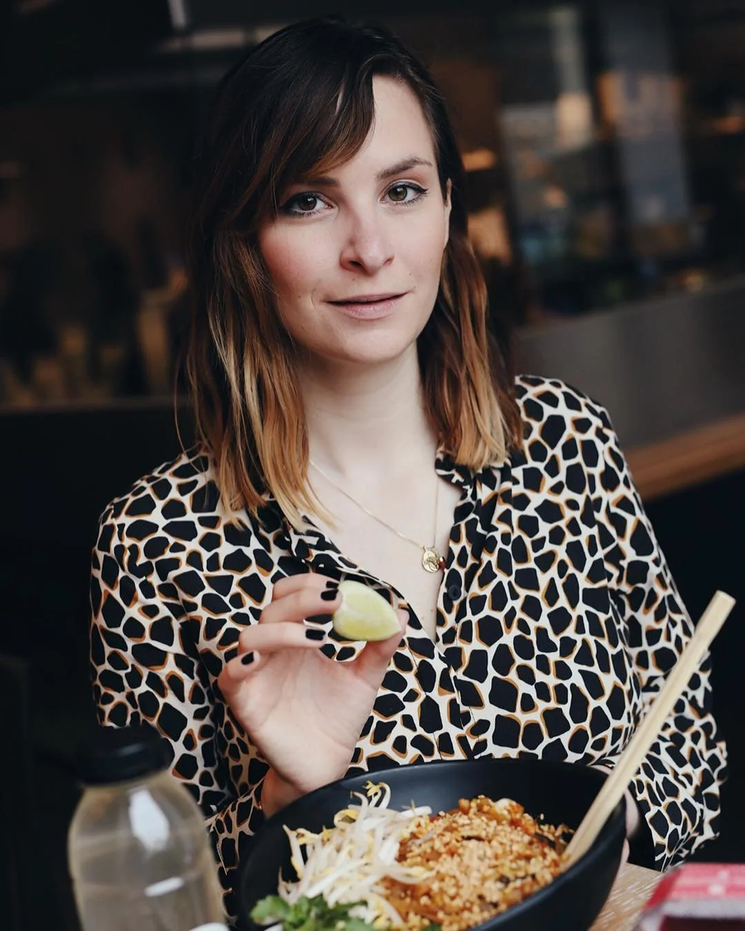 A woman with shoulder-length brown hair and light skin sitting at a restaurant table, holding a lime wedge, with a bowl of pad thai in front of her.