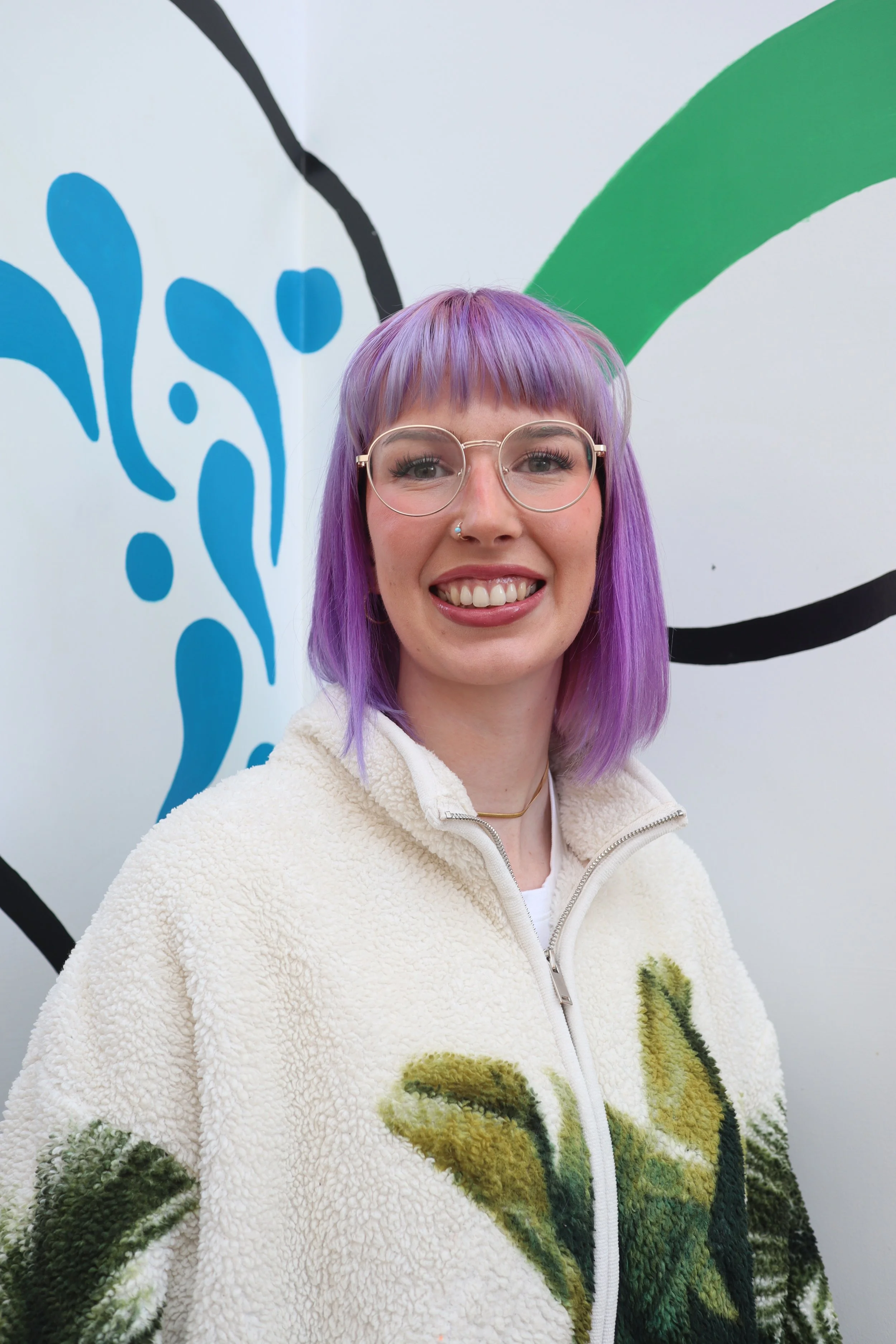 A smiling person with purple hair and glasses stands against a colourful wall mural with blue and green abstract shapes, wearing a fluffy jacket.