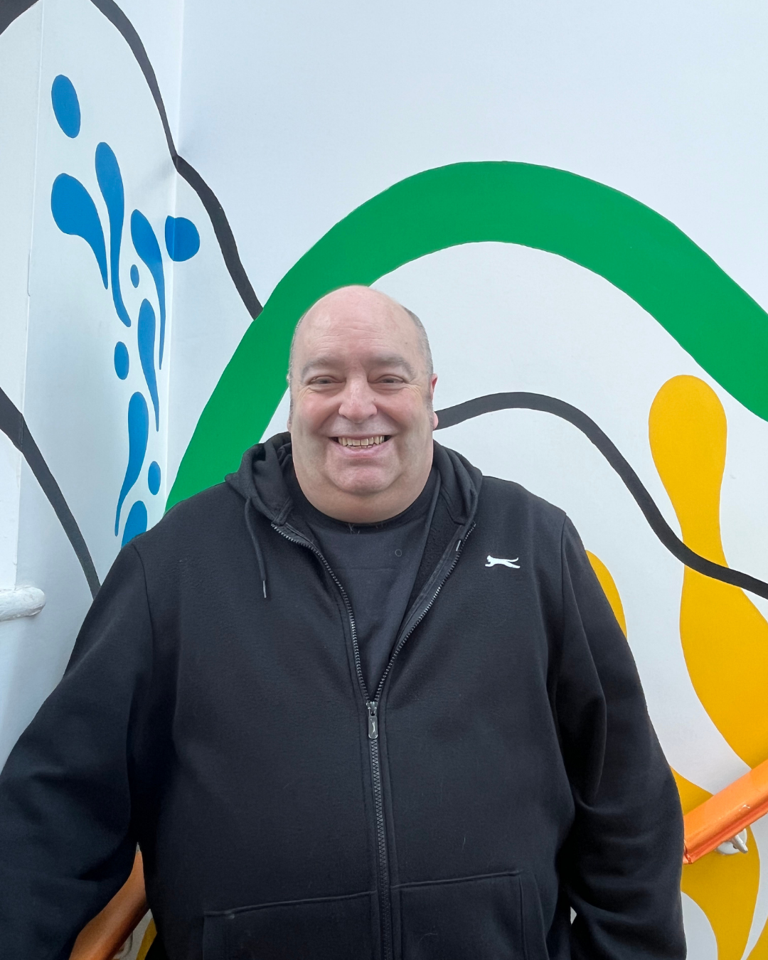 A man in a black hoodie stands smiling in front of a coloruful mural with abstract green, yellow, and blue shapes, conveying a cheerful and vibrant atmosphere.