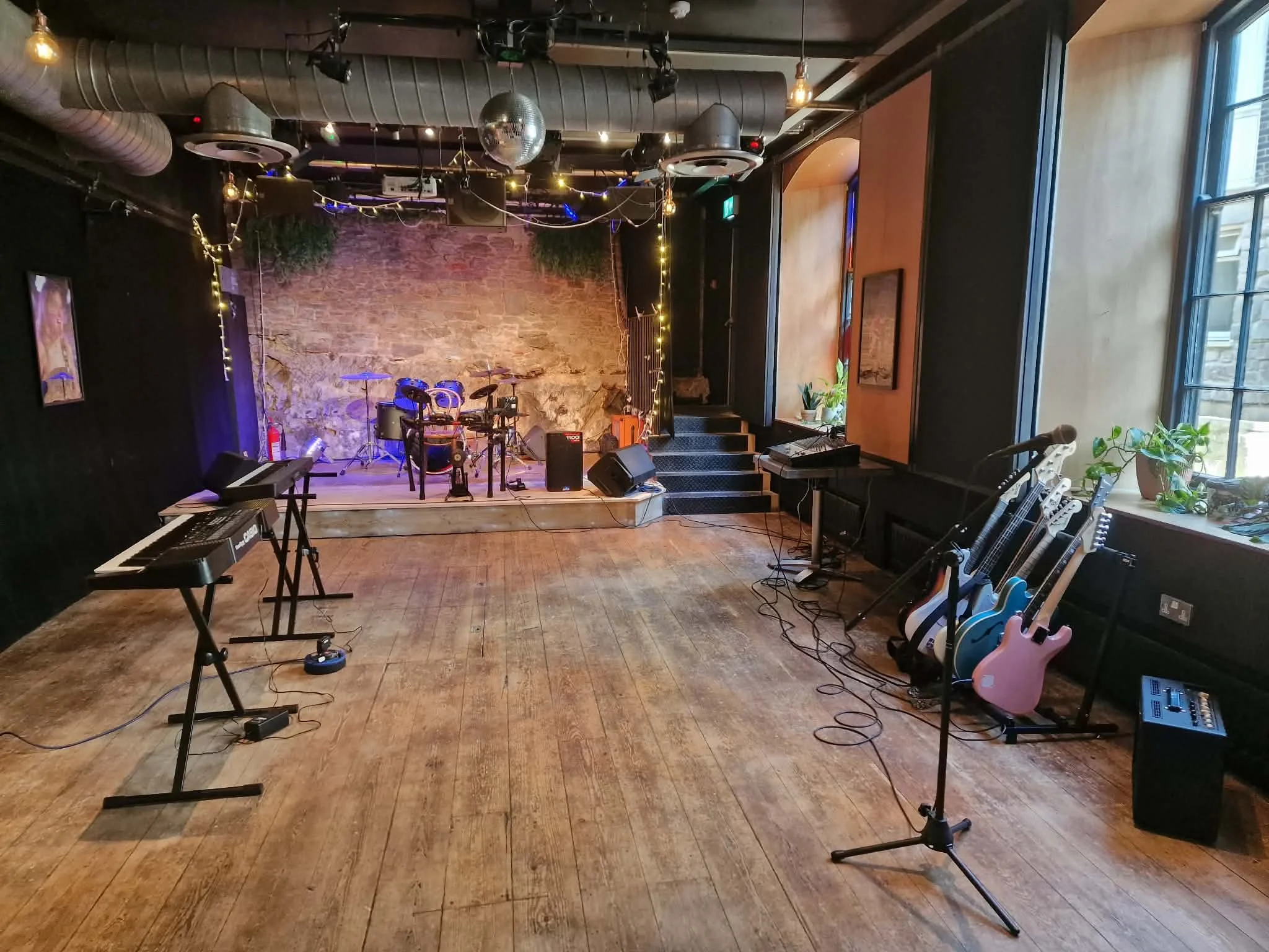 A cozy, dimly-lit restaurant with a small cabaret stage framed by red curtains and purple lighting. Tables with colourful chairs are set with menus, creating an inviting atmosphere.