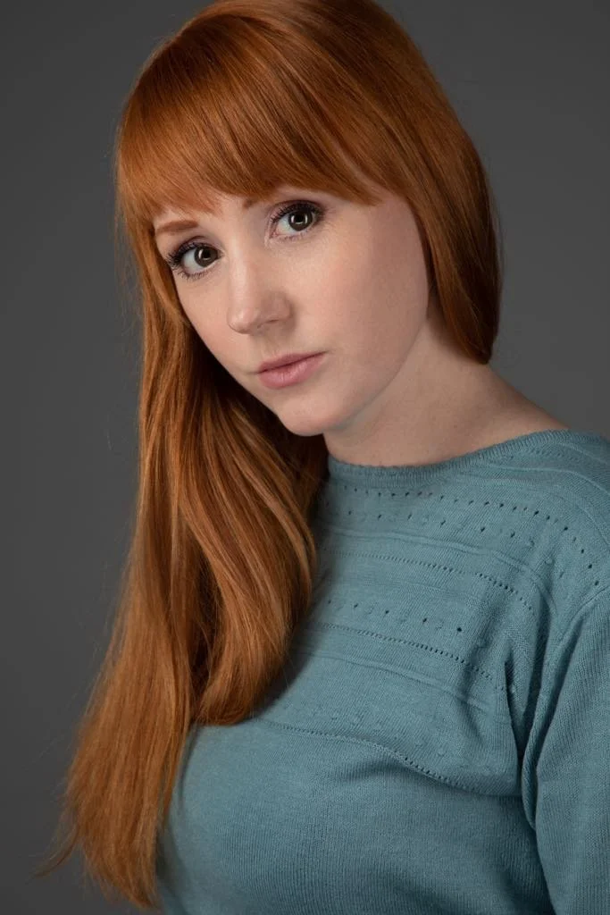A woman with long red hair and bangs, wearing a teal sweater, gazes softly at the camera against a grey background, conveying a serene mood.