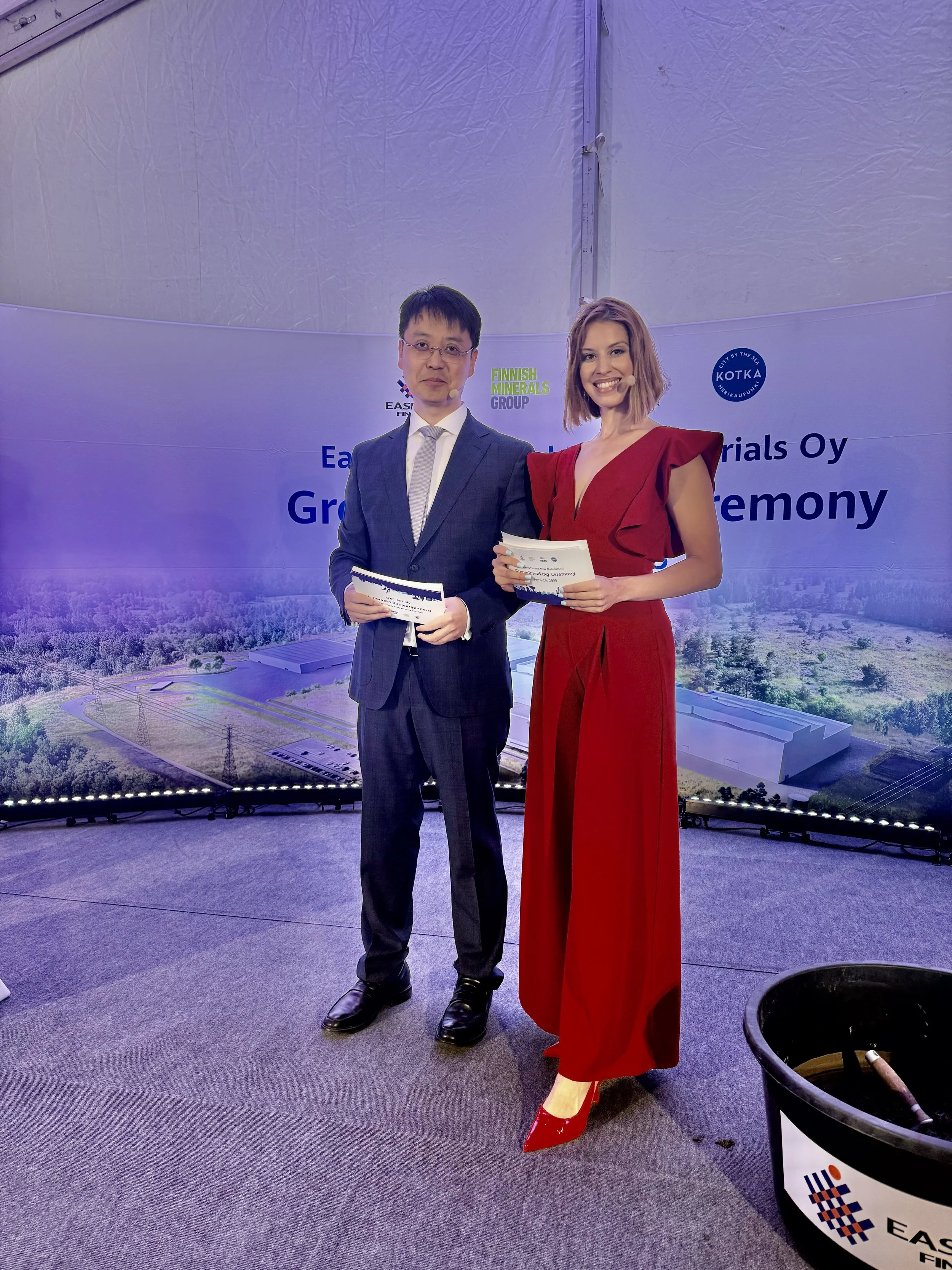 A man and woman in formal attire on stage during an award ceremony, holding certificates. The backdrop displays logos and the text "East Fin Materials Oy Group Ceremony."