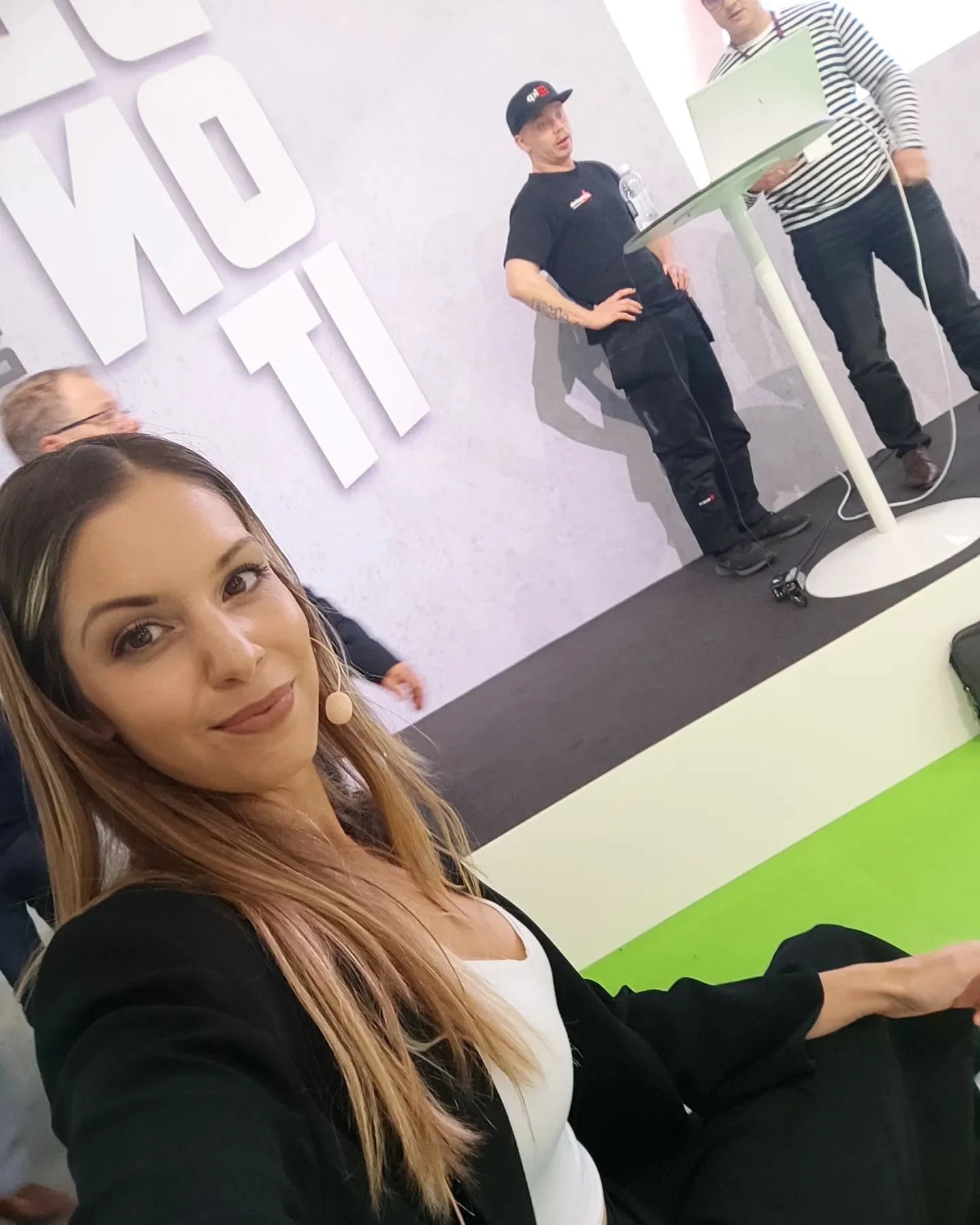 A woman taking a selfie at a conference or event, with a stage in the background showing two men standing near a computer and large letters on the wall.