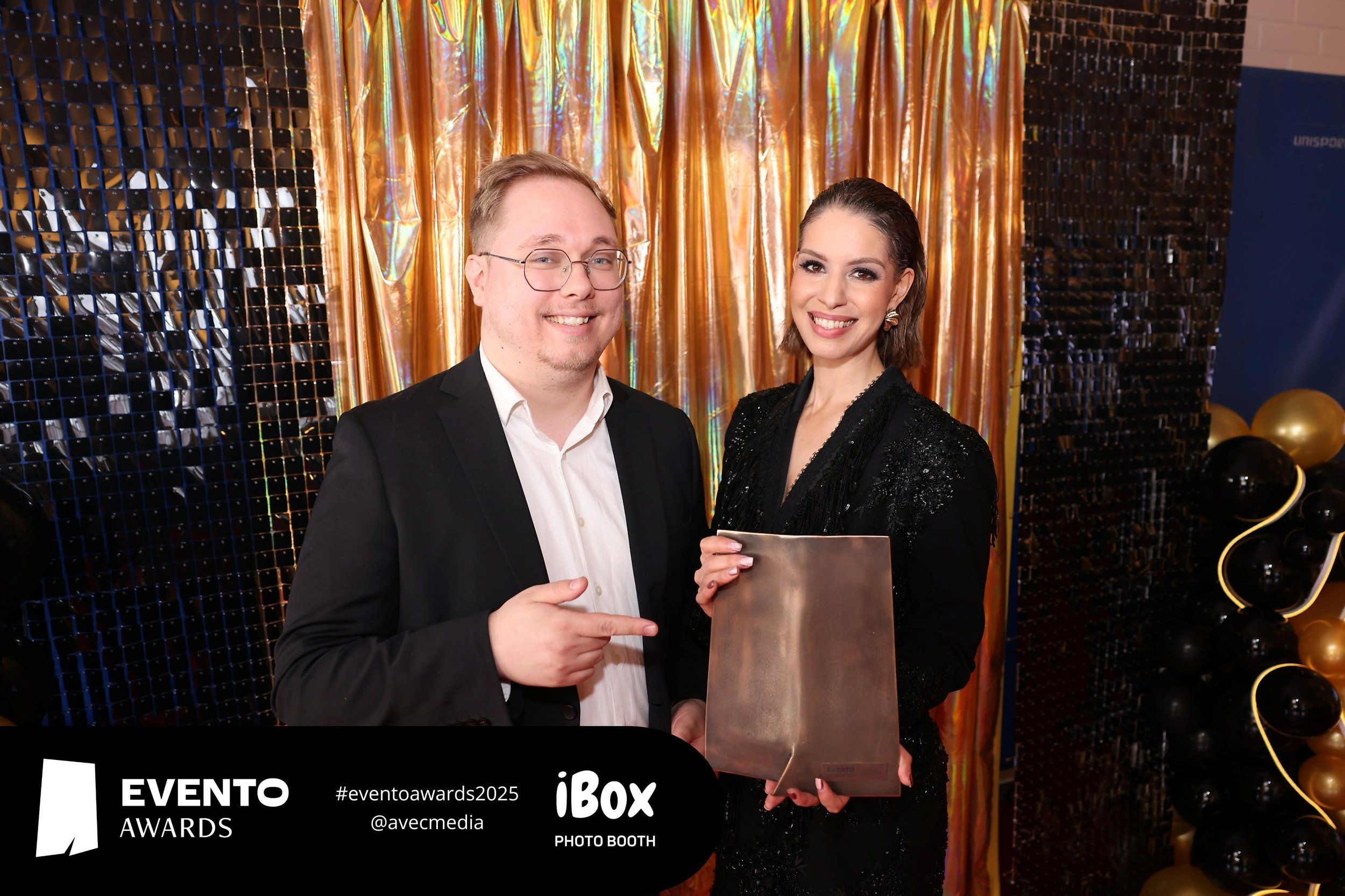 A man and a woman pose in front of a gold and black decorative backdrop at an award event. The man is wearing glasses and a black suit with a white shirt, pointing at the woman, who is holding a bronze-colored award and is dressed in a black, sequine