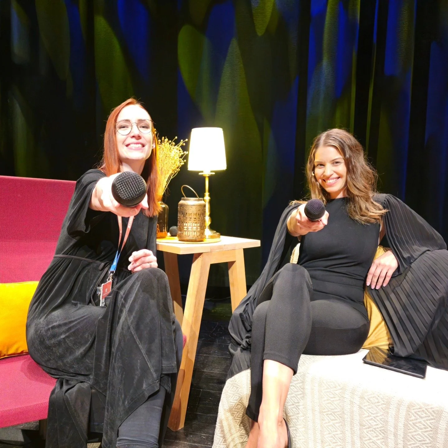 Two women sitting on a couch, smiling and holding microphones towards the camera, in a cozy, well-lit indoor setting with a side table, lamp, and decorative items in the background.
