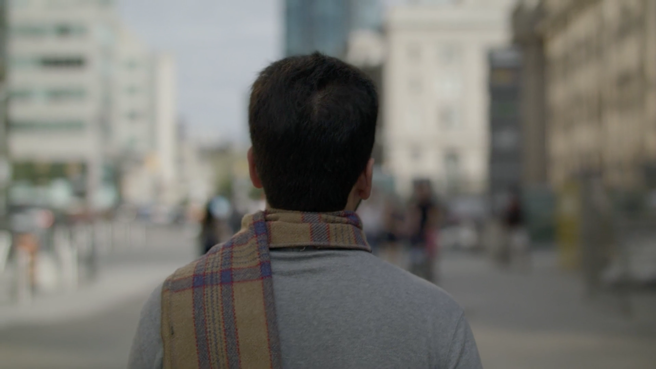 Un homme vu de dos qui marche dans une ville, portant un manteau gris et une écharpe à carreaux, avec des bâtiments et des personnes flous en arrière-plan.