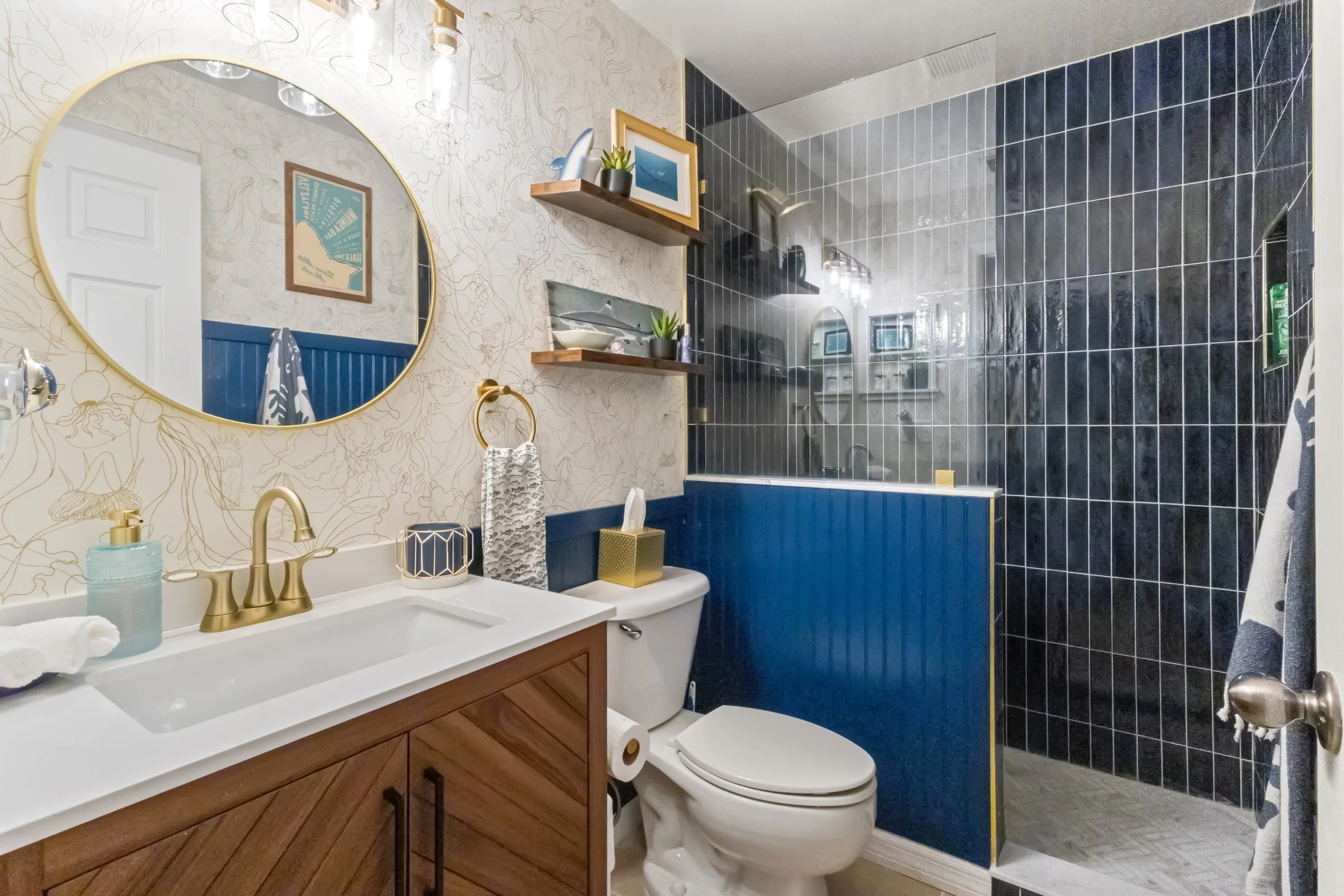 A modern bathroom with a wooden vanity, a gold faucet, and a round mirror. The walls are decorated with floral wallpaper and framed artwork. A toilet with a gold tissue box on top is next to the vanity. There are floating wooden shelves with plants a