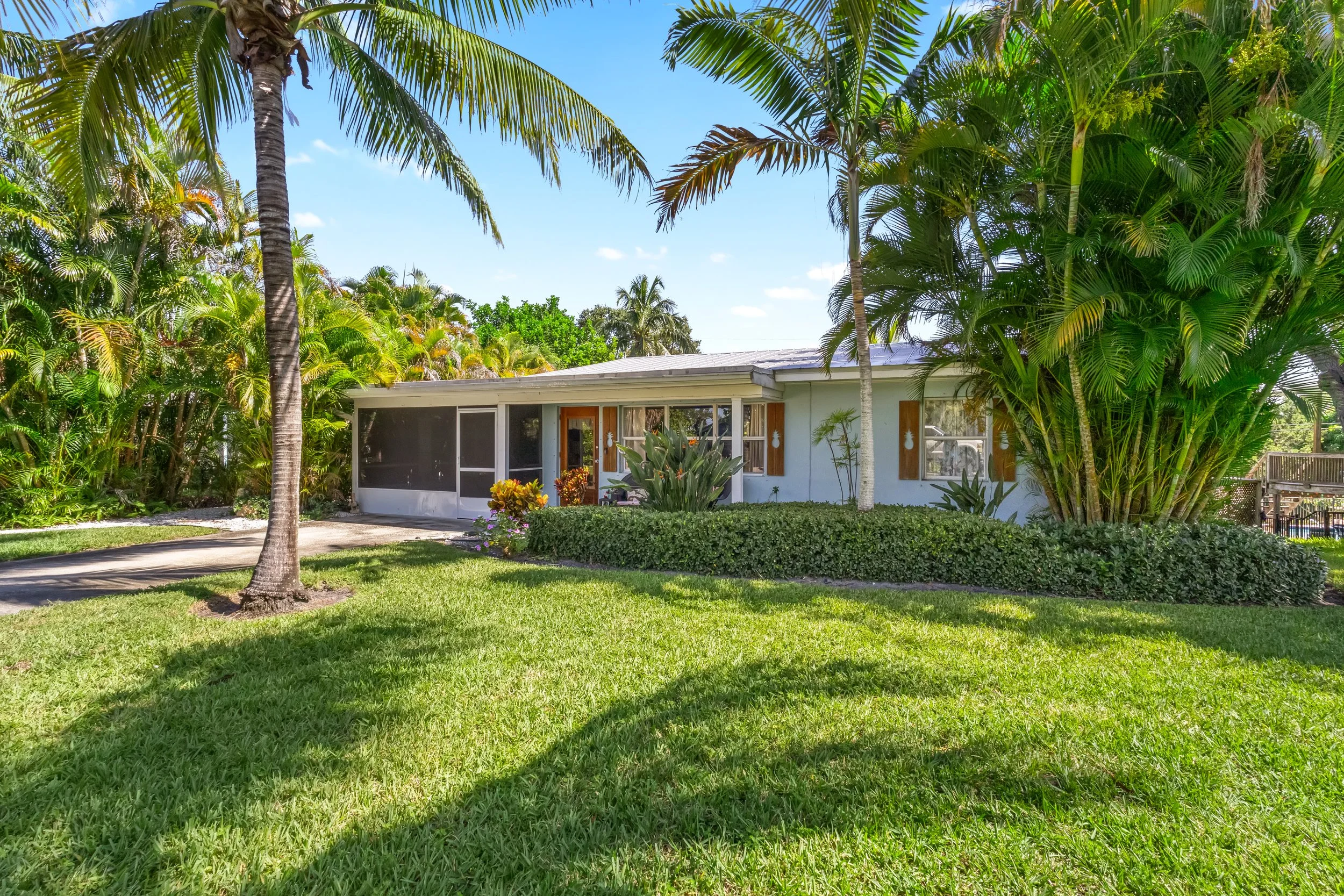 A single-story house in a tropical setting with lush green grass, palm trees, and dense tropical plants surrounding it, under a blue sky.