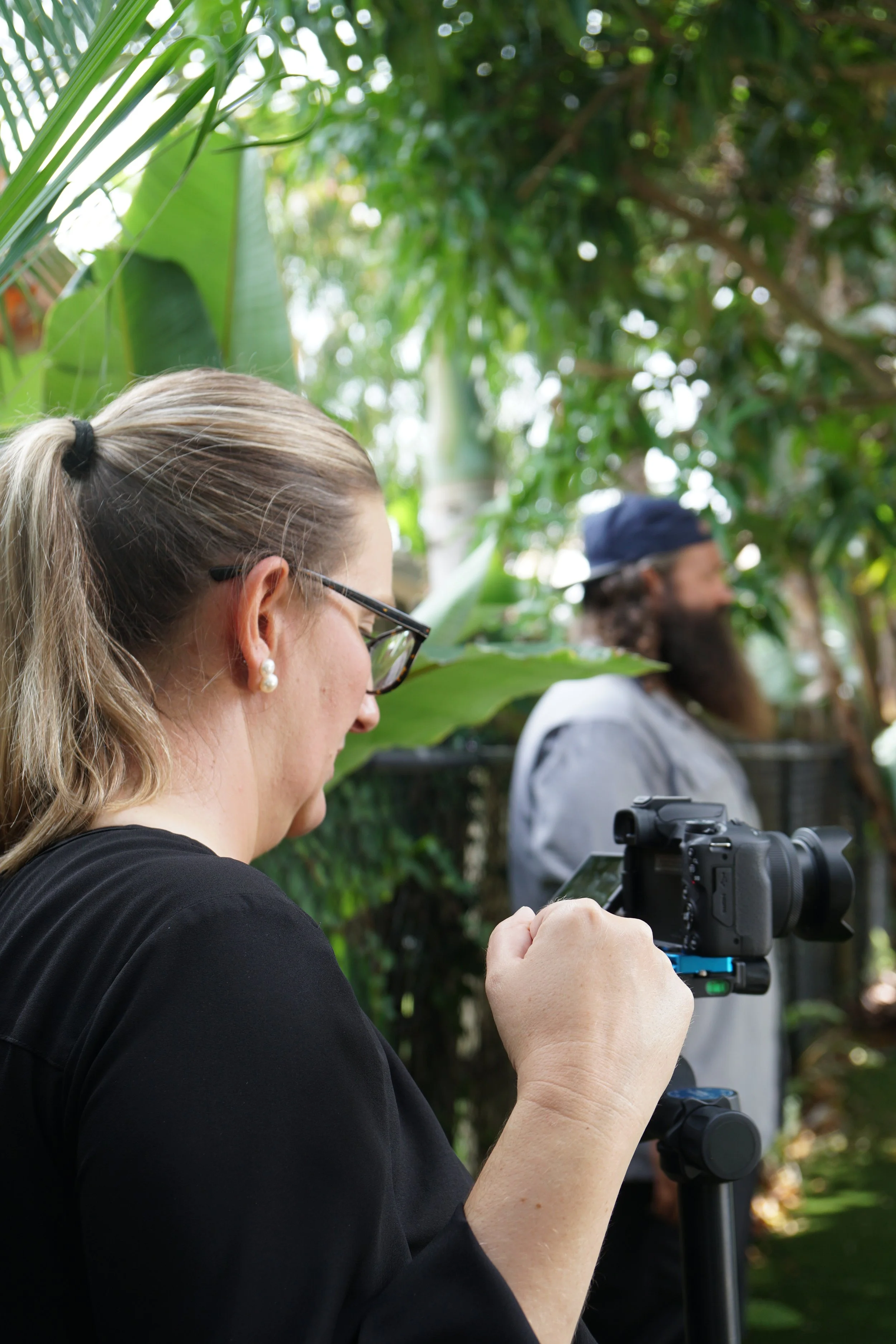 A woman in black shirt with glasses and pearl earrings filming or photographing with a camera on a tripod, in a lush green outdoor setting. A man with a beard, gray shirt, and cap is blurred in the background.