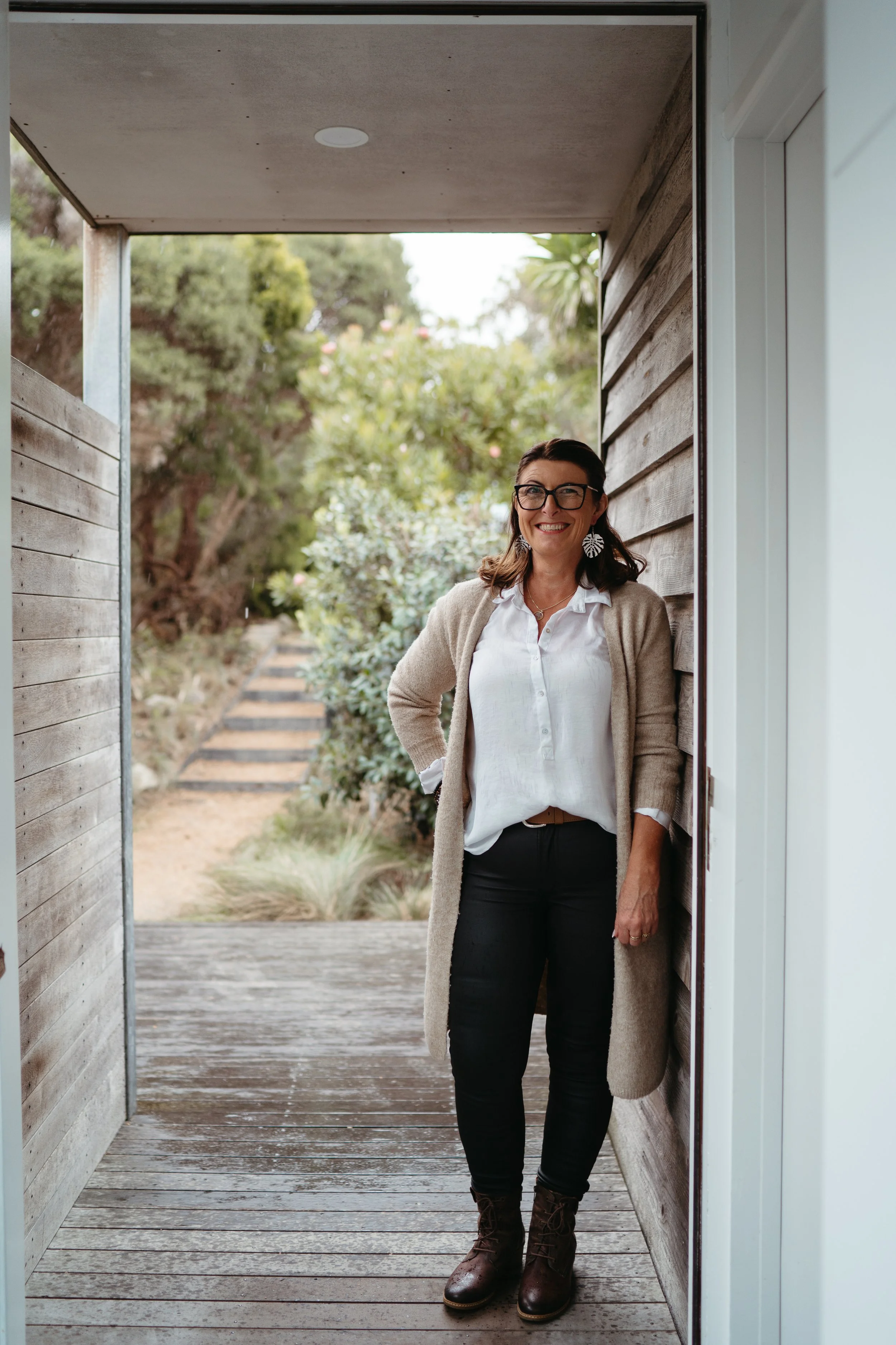 A woman standing on a wooden porch, smiling, with greenery in the background.