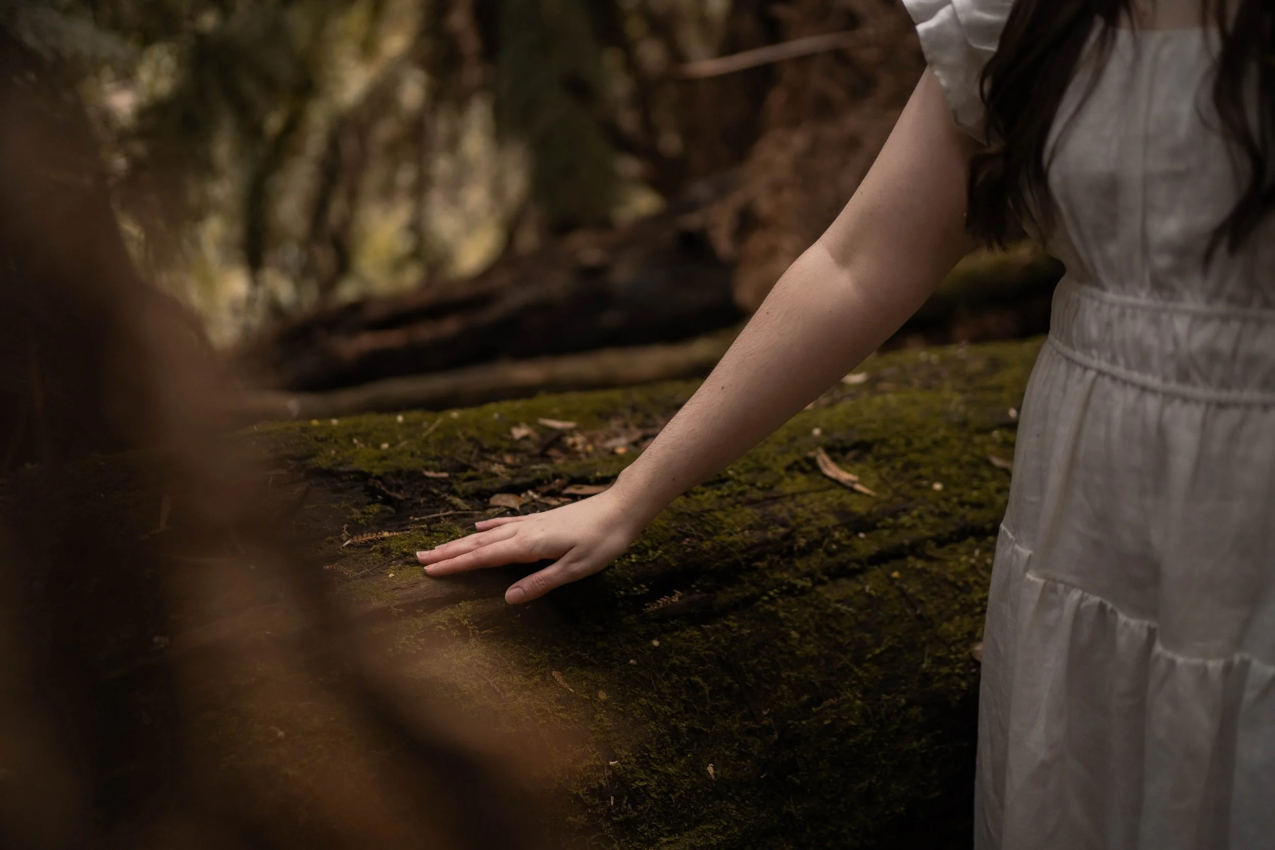 A person in a white dress with their hand on a moss-covered fallen log in a forest.