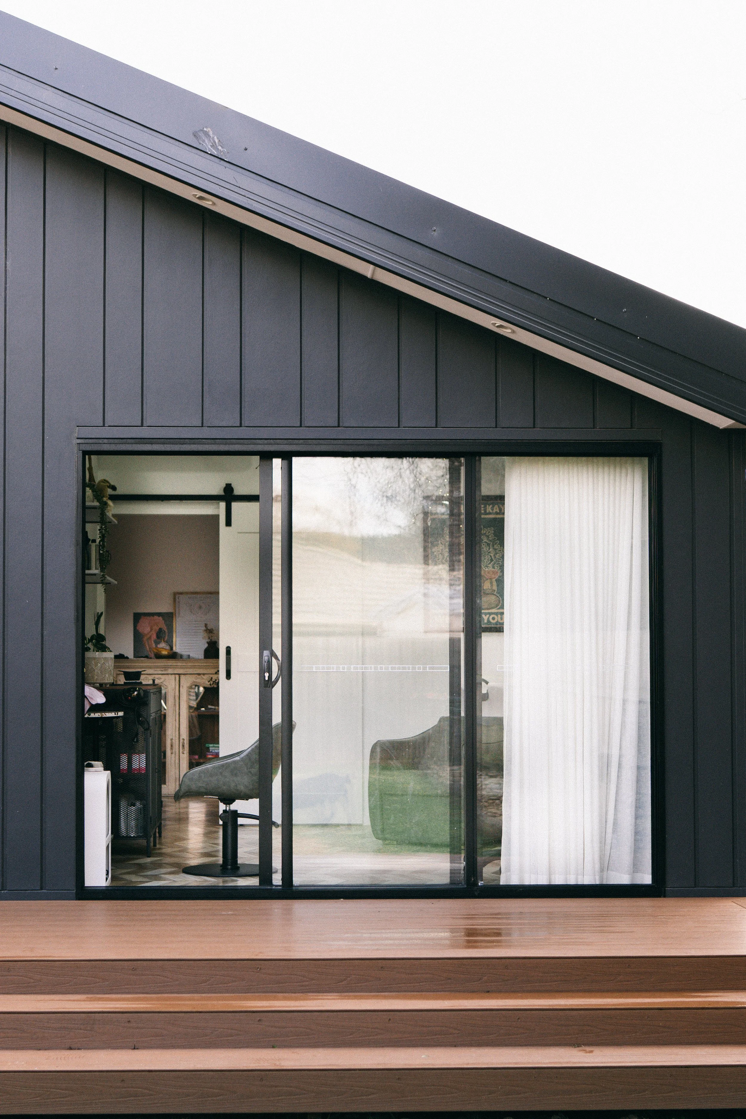 View of a modern house with a sliding glass door, dark vertical siding, and a wooden deck, with a glimpse into a home interior.