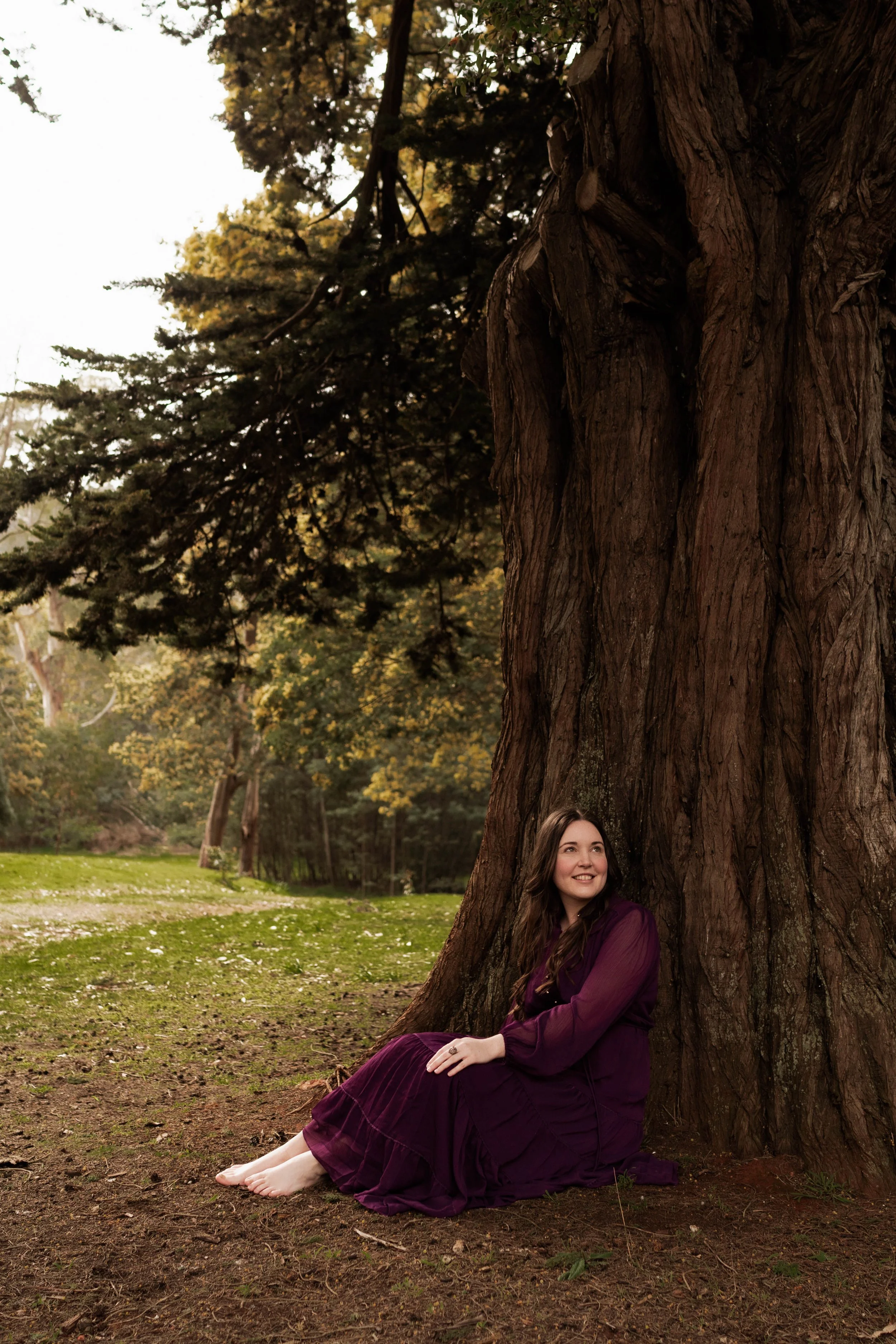 A woman in a purple dress sitting barefoot on the ground, leaning against a large tree in a park with green grass and trees in the background.