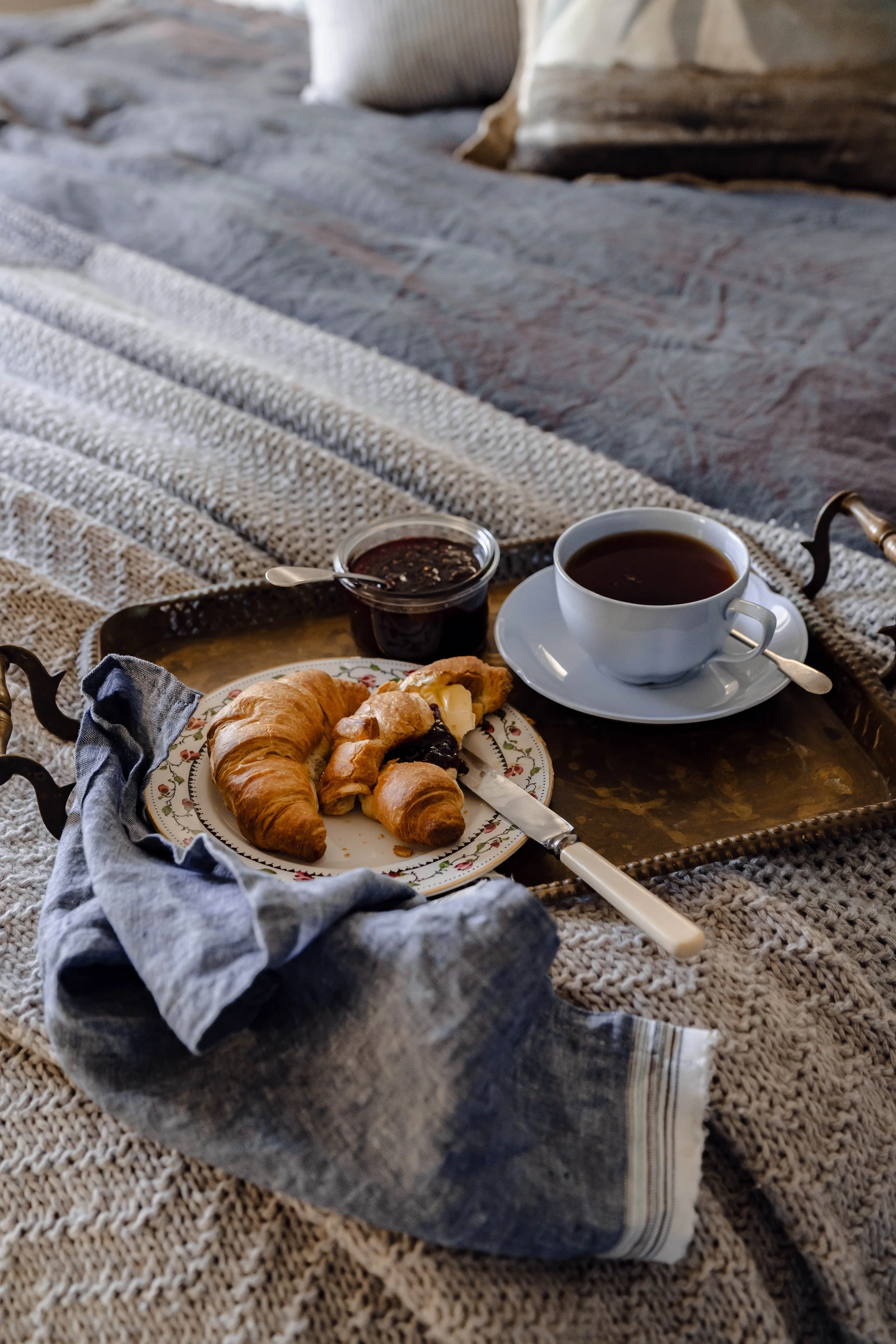 A breakfast tray on a bed with croissants, jam, a cup of coffee, and a knife.