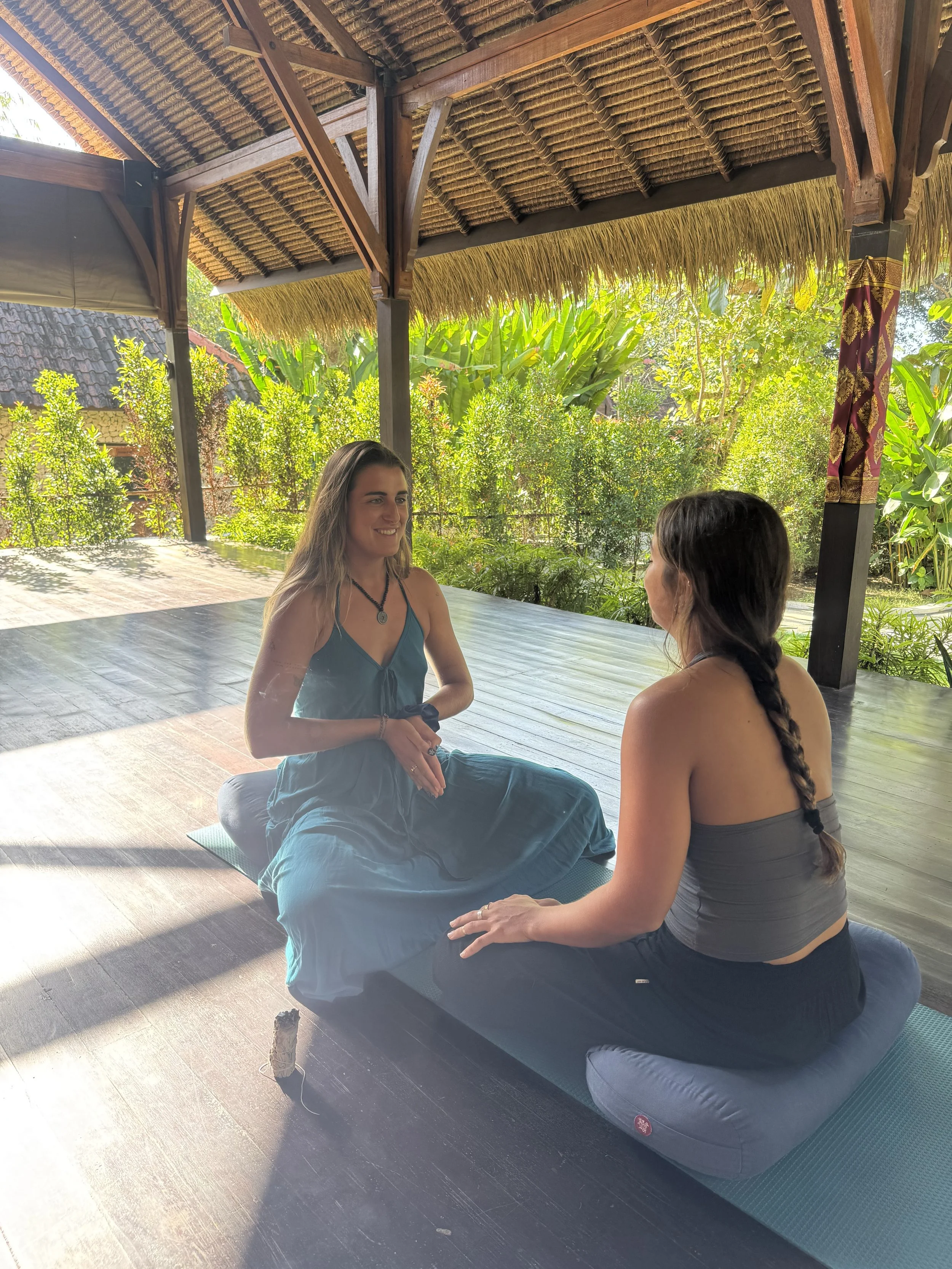 Dos mujeres practicando yoga o meditación en un espacio abierto y natural, con techo de paja y rodeadas de vegetación verde.