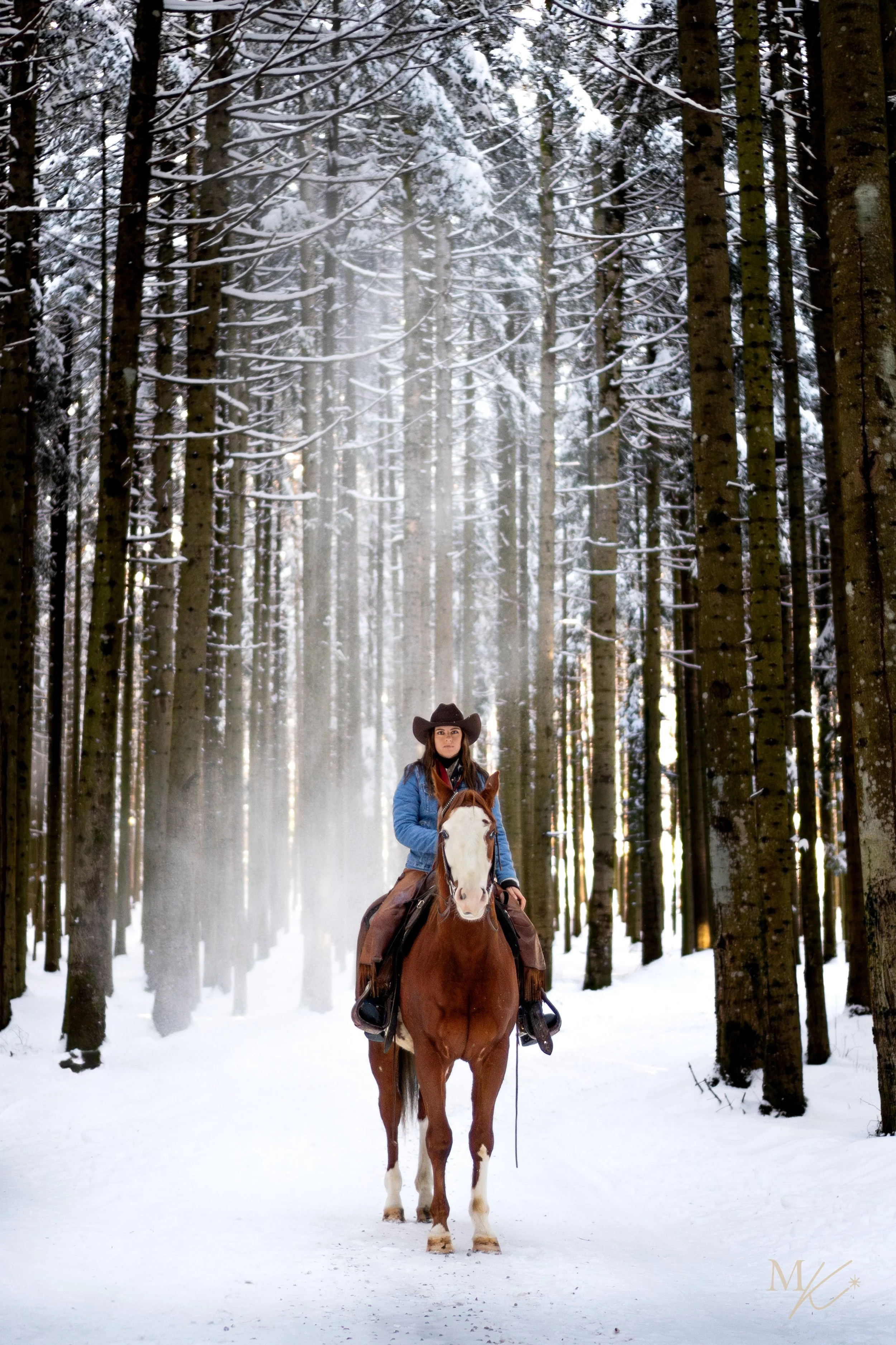 A woman wearing a cowboy hat and blue jacket riding a brown and white horse through a snowy forest.