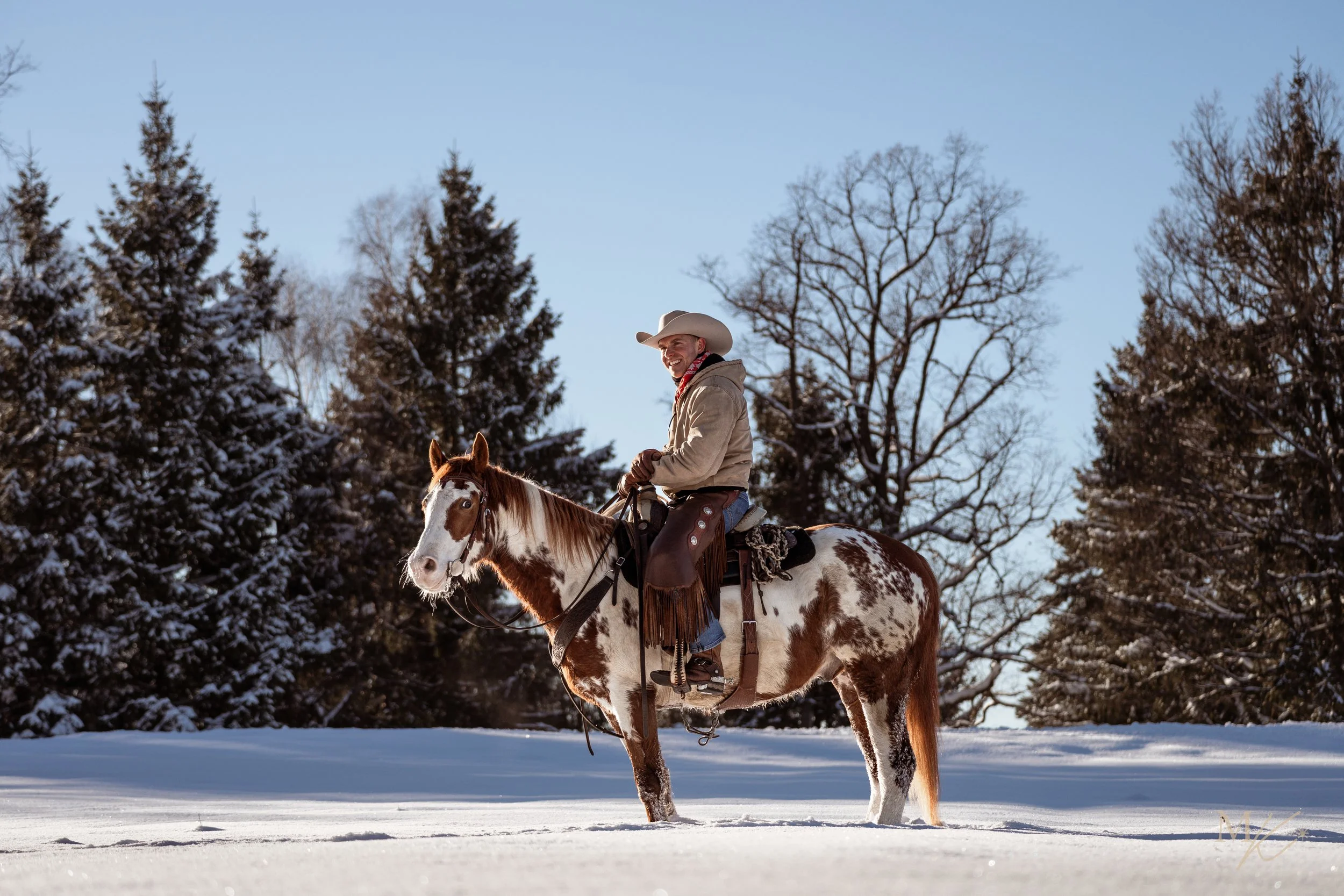 A man riding a paint horse in a snowy landscape with pine trees and leafless trees in the background.