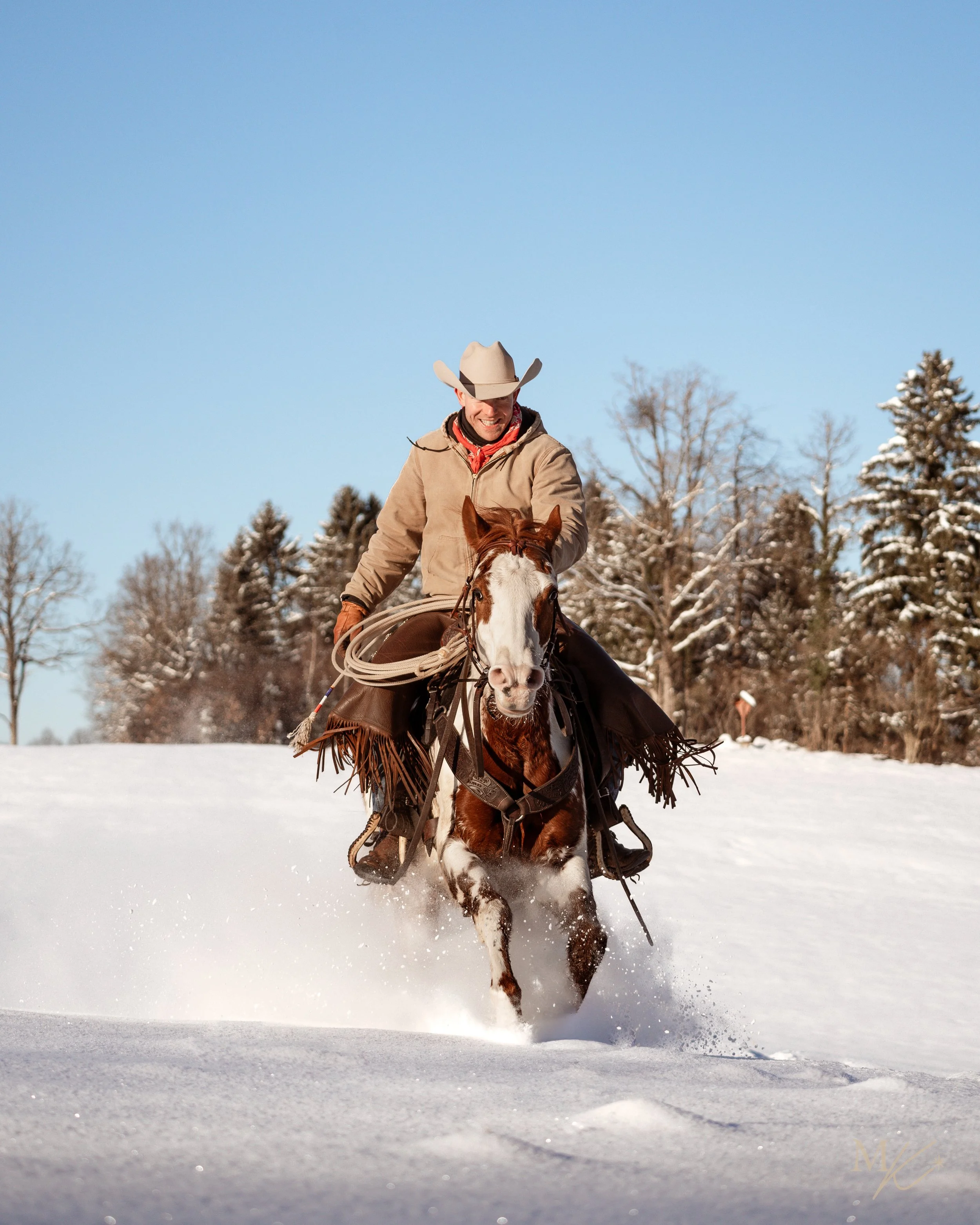 A cowboy riding a horse through snow on a sunny day with trees in the background.