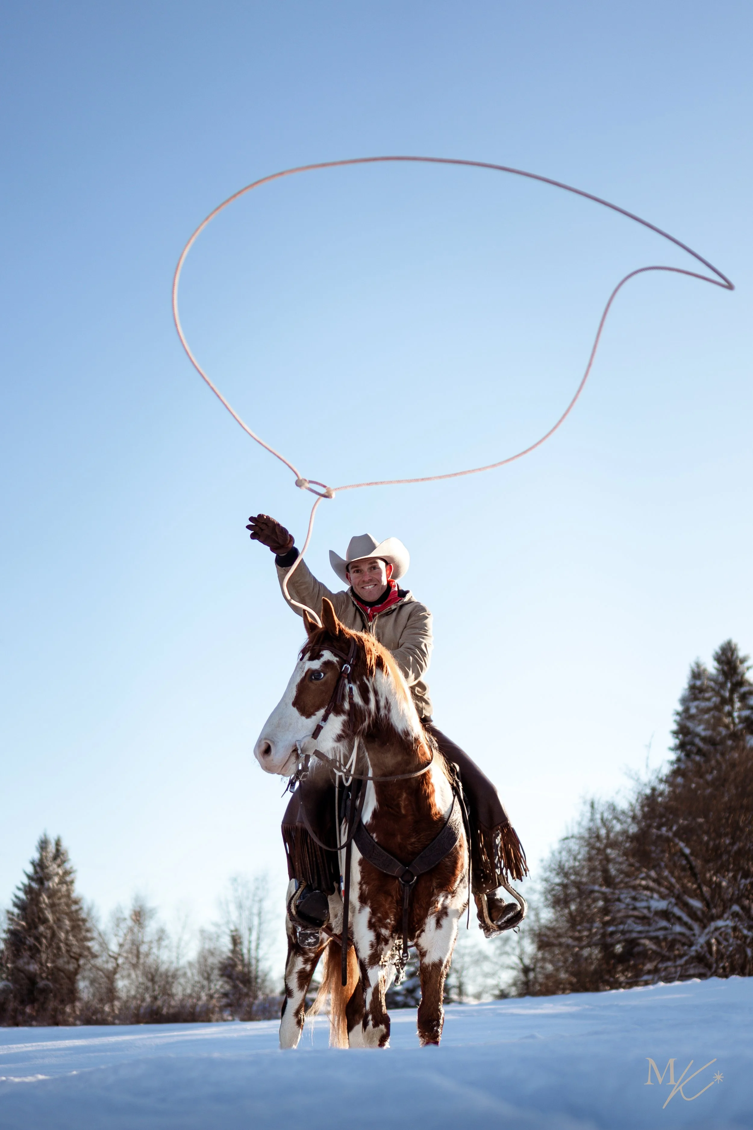 A cowboy riding a horse on a snowy landscape, smiling and swinging a lasso in the air during daytime.