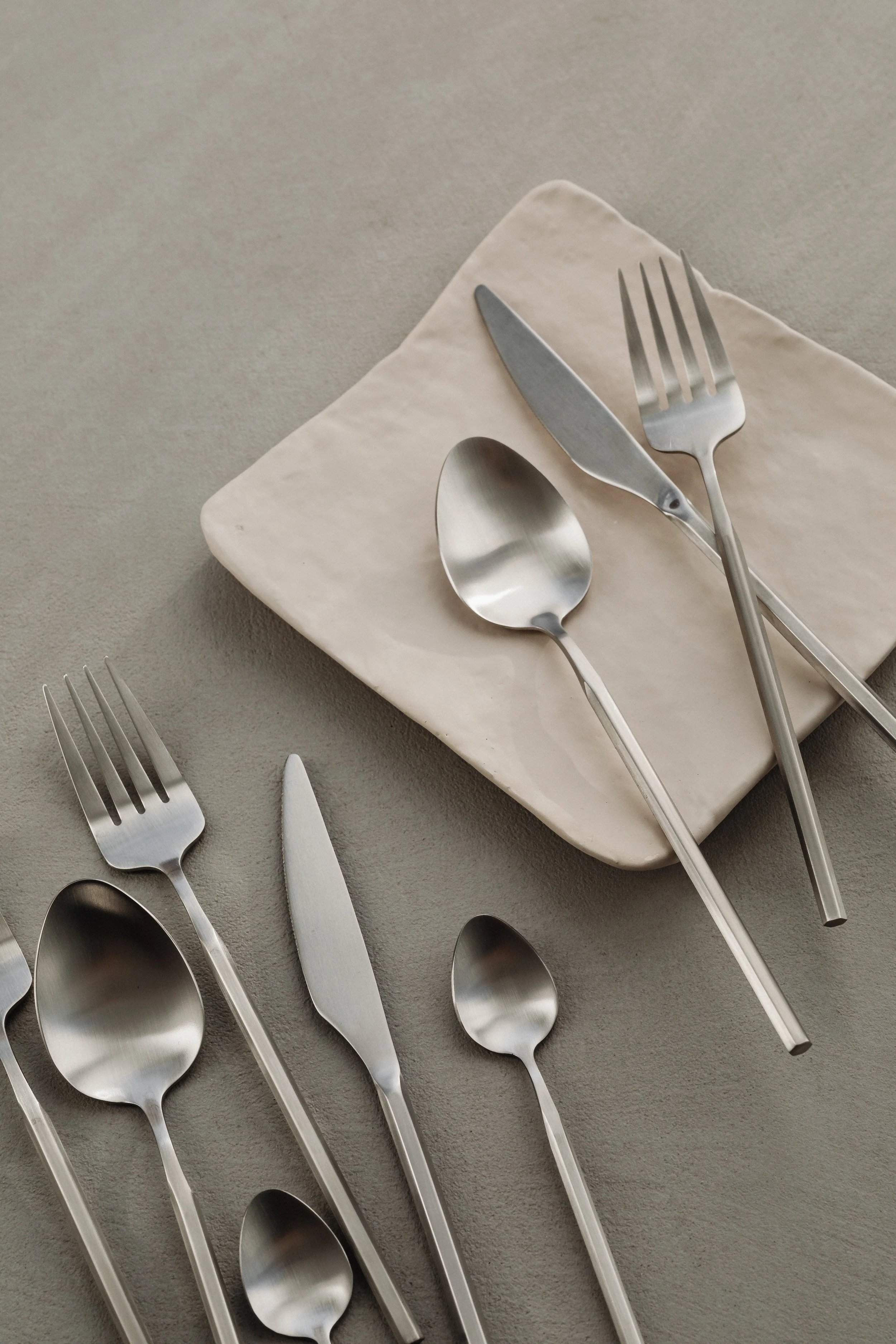 A set of stainless steel forks, knives, and spoons arranged on a light gray surface, with some utensils placed on a white rectangular plate.