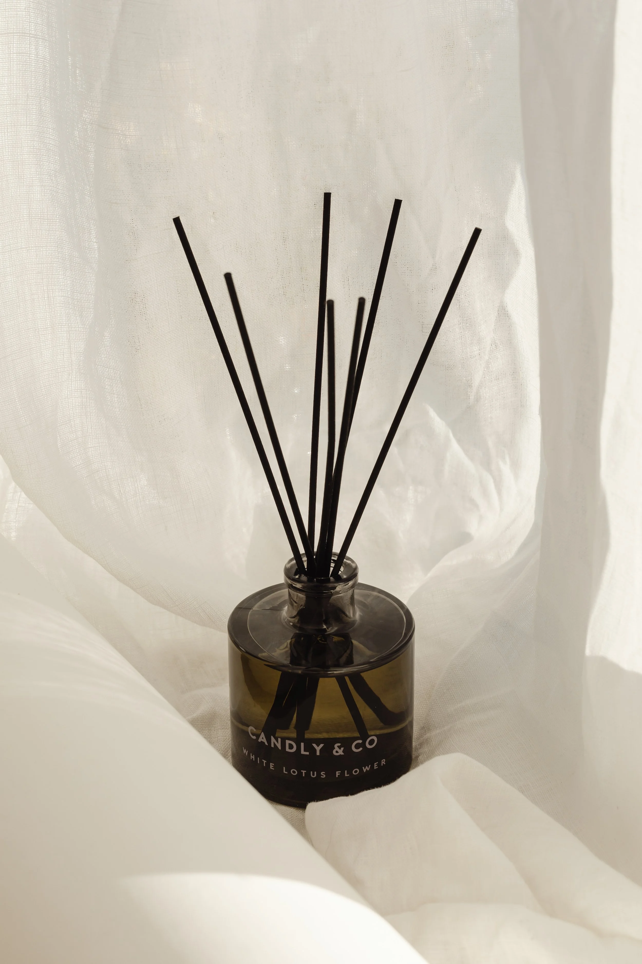 A dark glass diffuser bottle labeled 'Candly & Co White Lotus Flower' with black reed sticks inside, set against sheer white curtains.