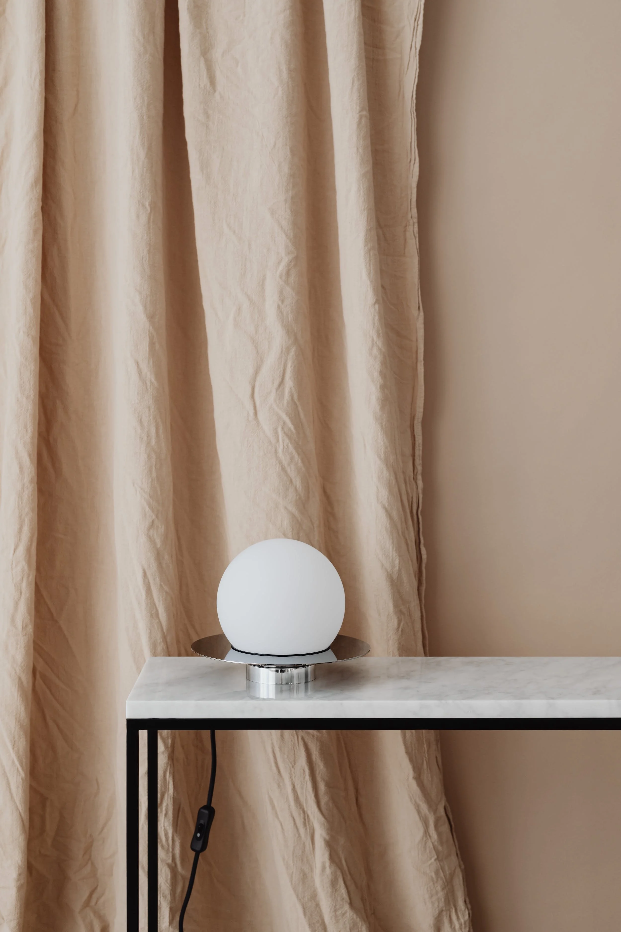 A modern white spherical lamp on a black and white marble side table with a beige curtain in the background.