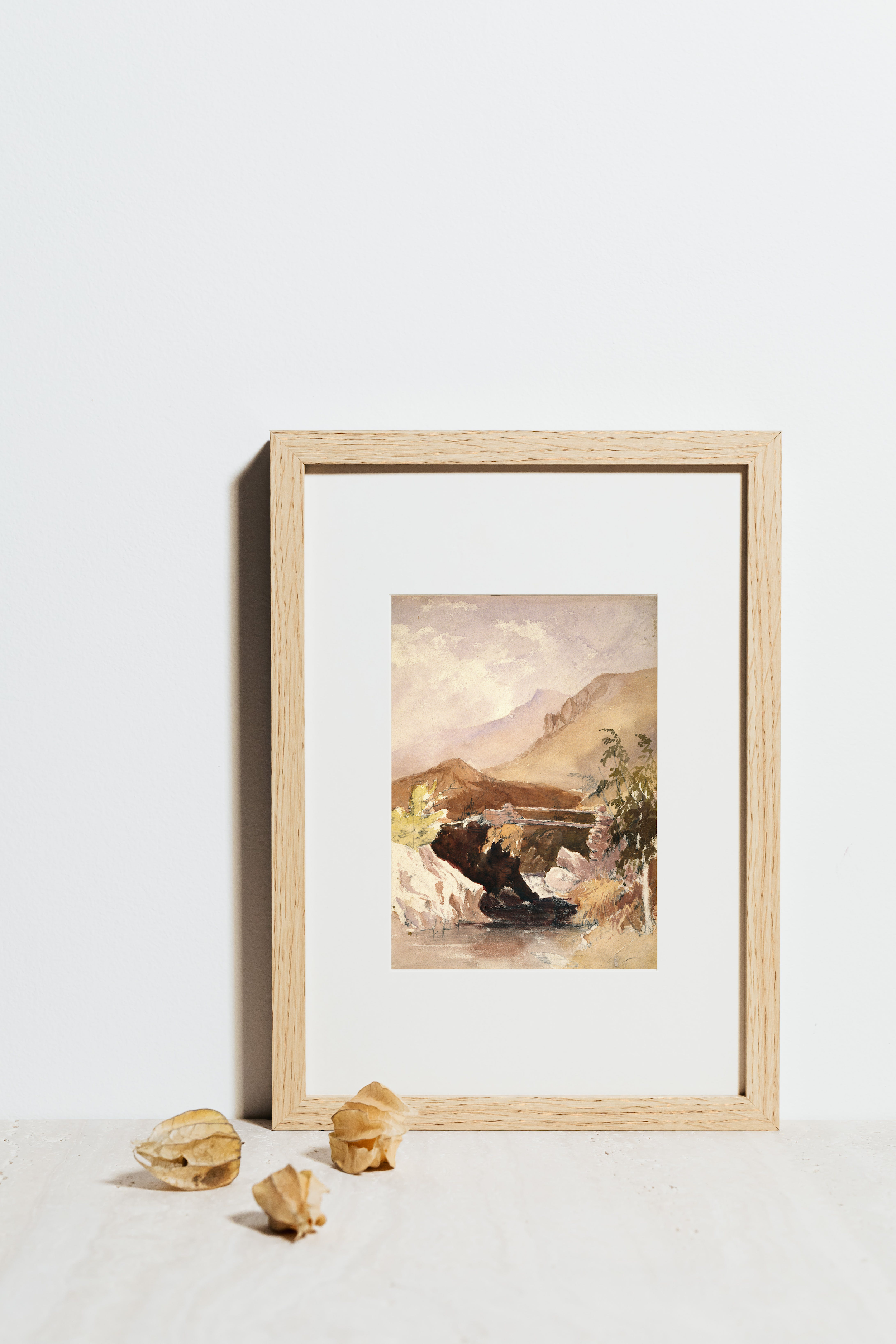 Framed landscape painting of mountains and a river, leaning against a white wall, with three dried leaves on the surface in front.