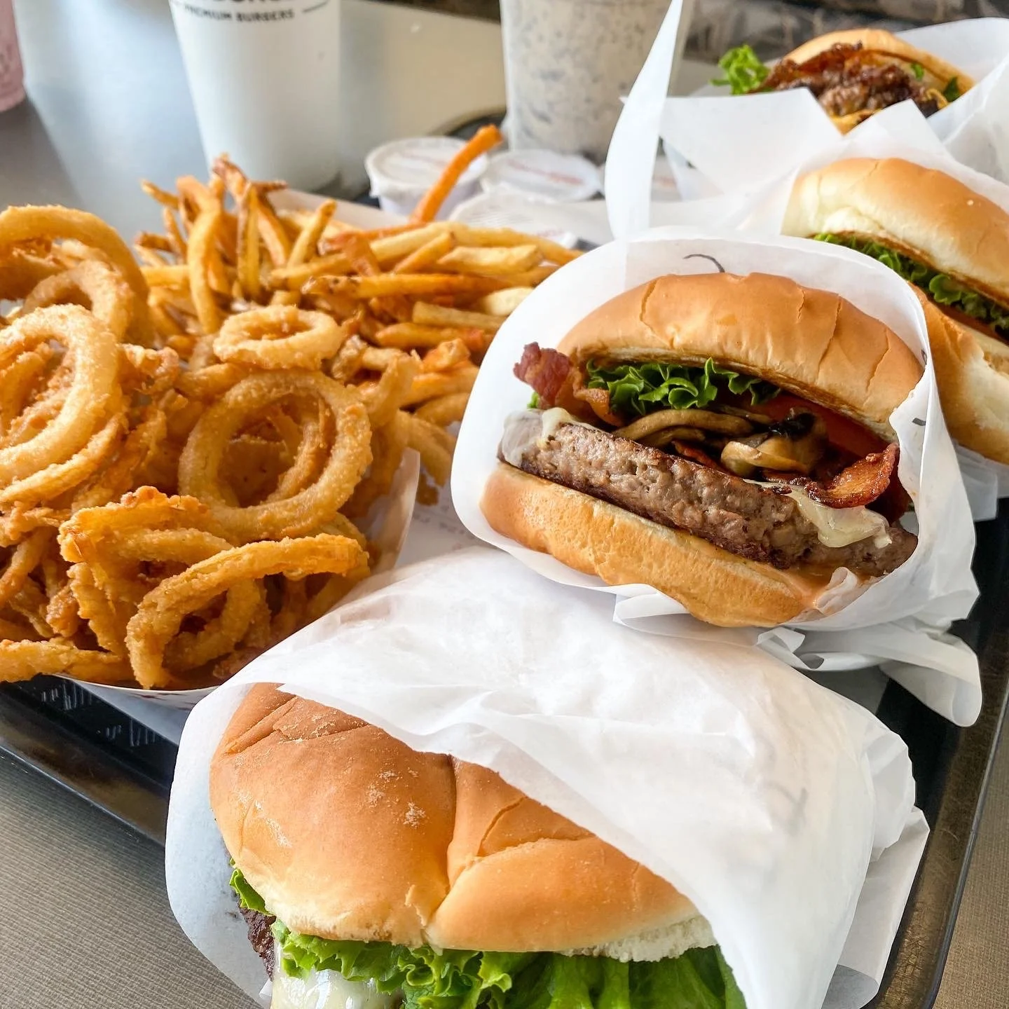 A tray with onion rings, French fries, and two cheeseburgers, one unwrapped and one partially wrapped, served with lettuce, tomato, bacon, and mushrooms.