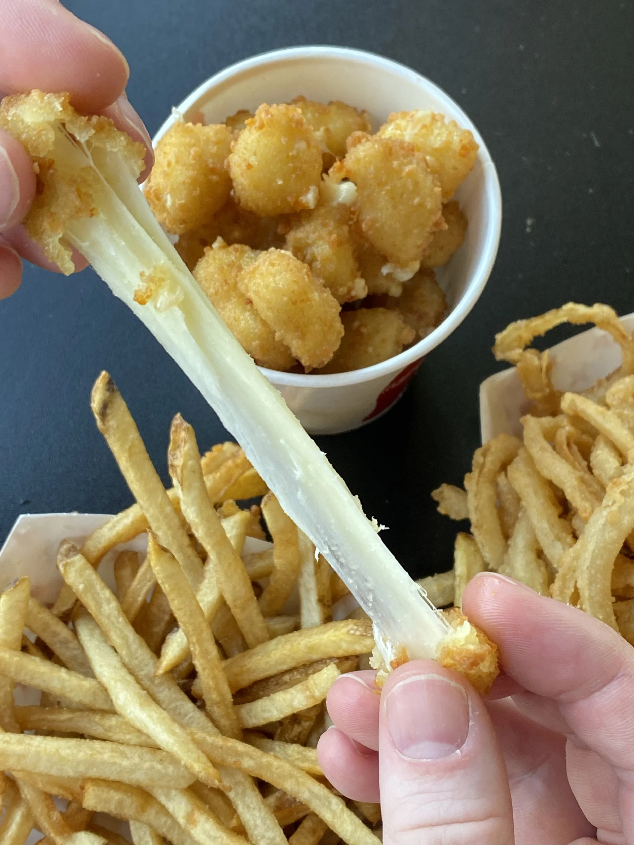 Person peeling a mozzarella cheese stick over a tray of French fries, with a cup of fried chicken nuggets in the background.