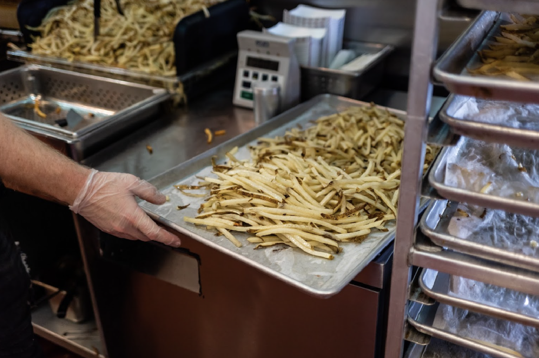 A person wearing a disposable glove is holding a tray of cooked French fries in a commercial kitchen or restaurant.
