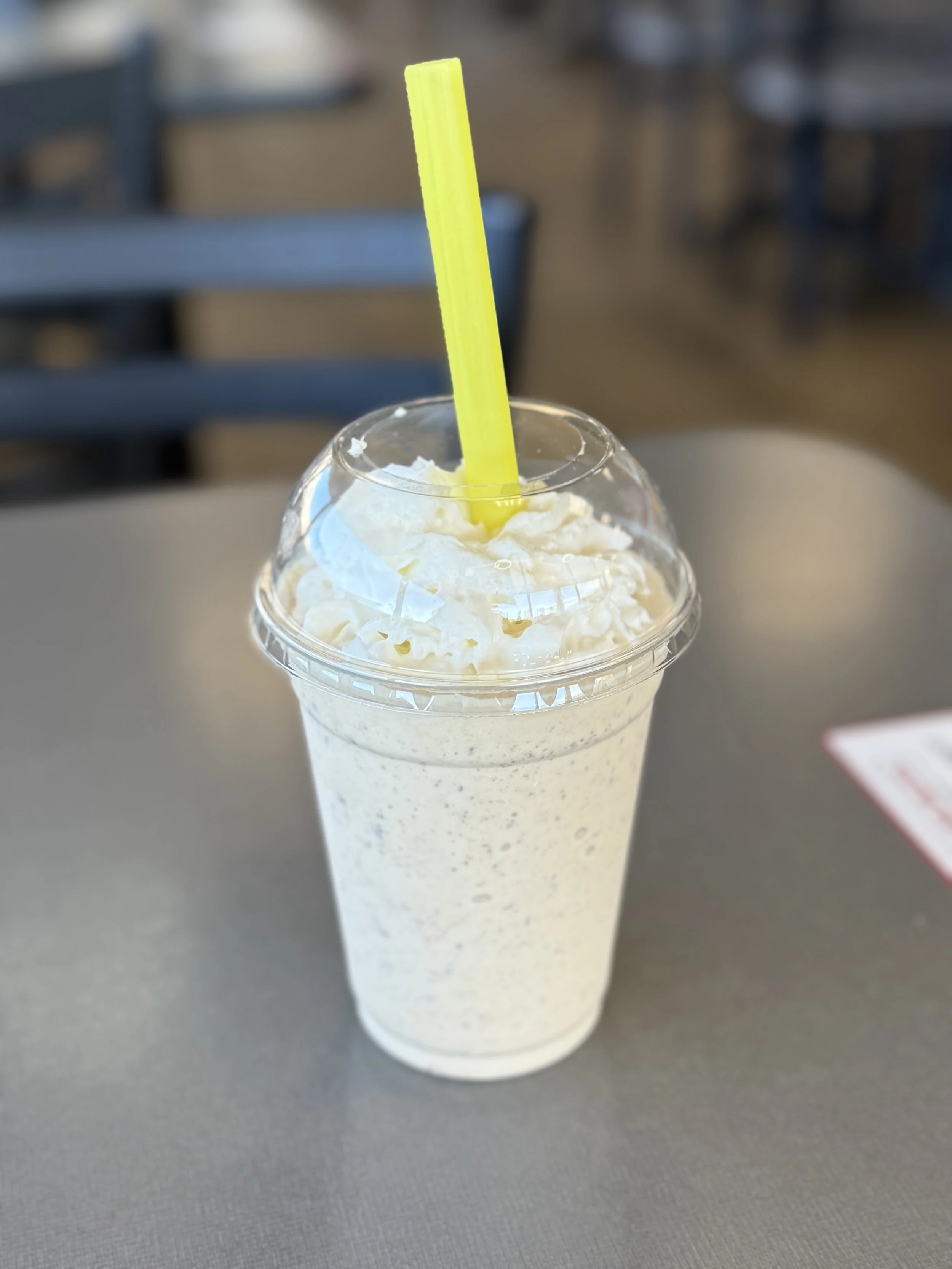 A vanilla milkshake topped with whipped cream and served with a yellow straw in a clear plastic cup, on a gray table.