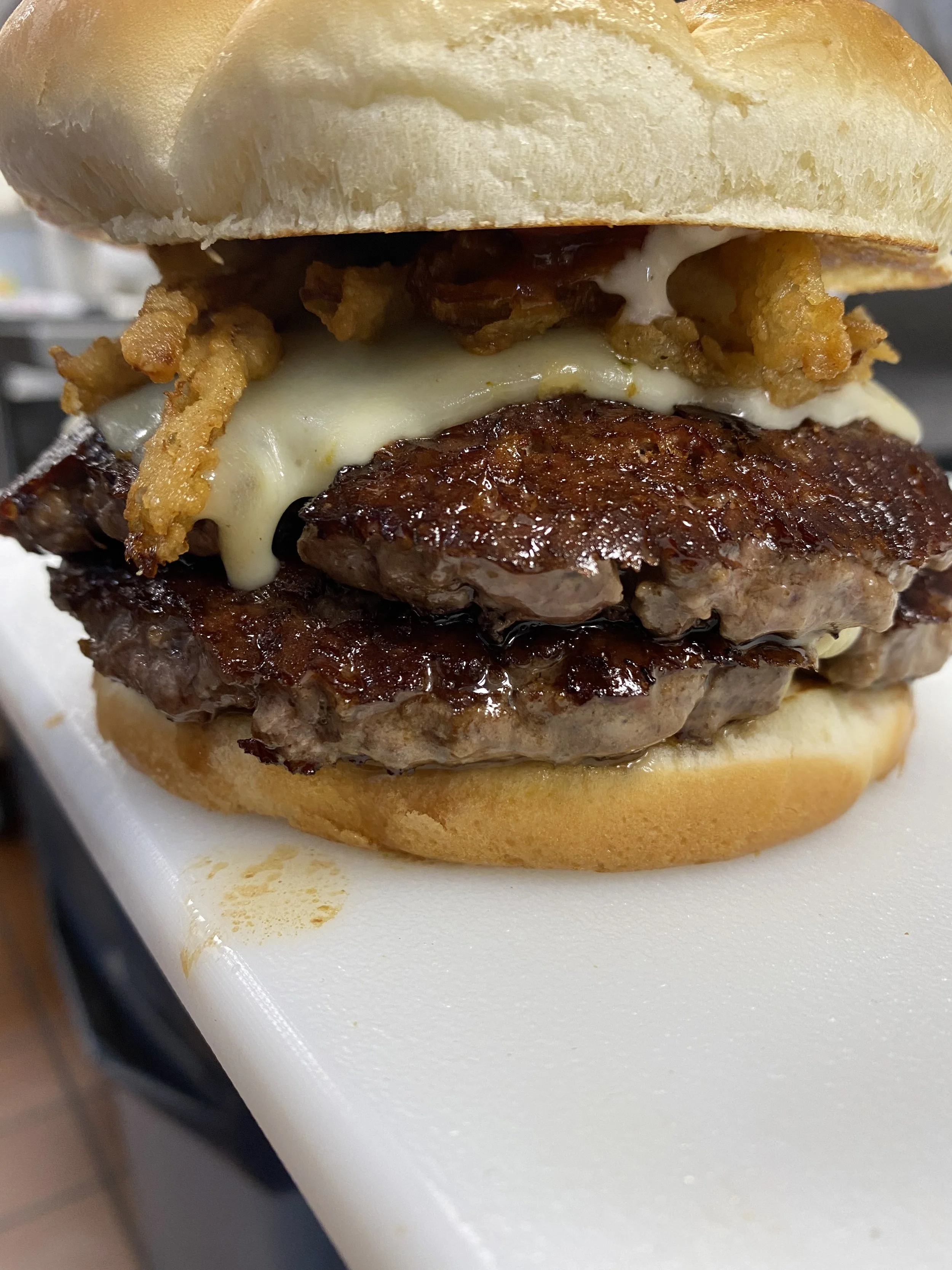A close-up of a cheeseburger with bacon, fried onion strings, melted cheese, grilled beef patty, lettuce, and a bun, placed on a white surface.