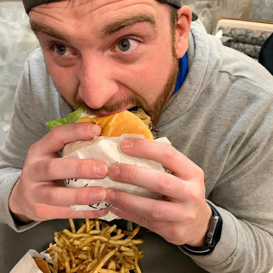 A man in a gray hoodie is about to eat a cheeseburger with lettuce, cheese, and meat, holding it with both hands. There are French fries on the table in front of him.
