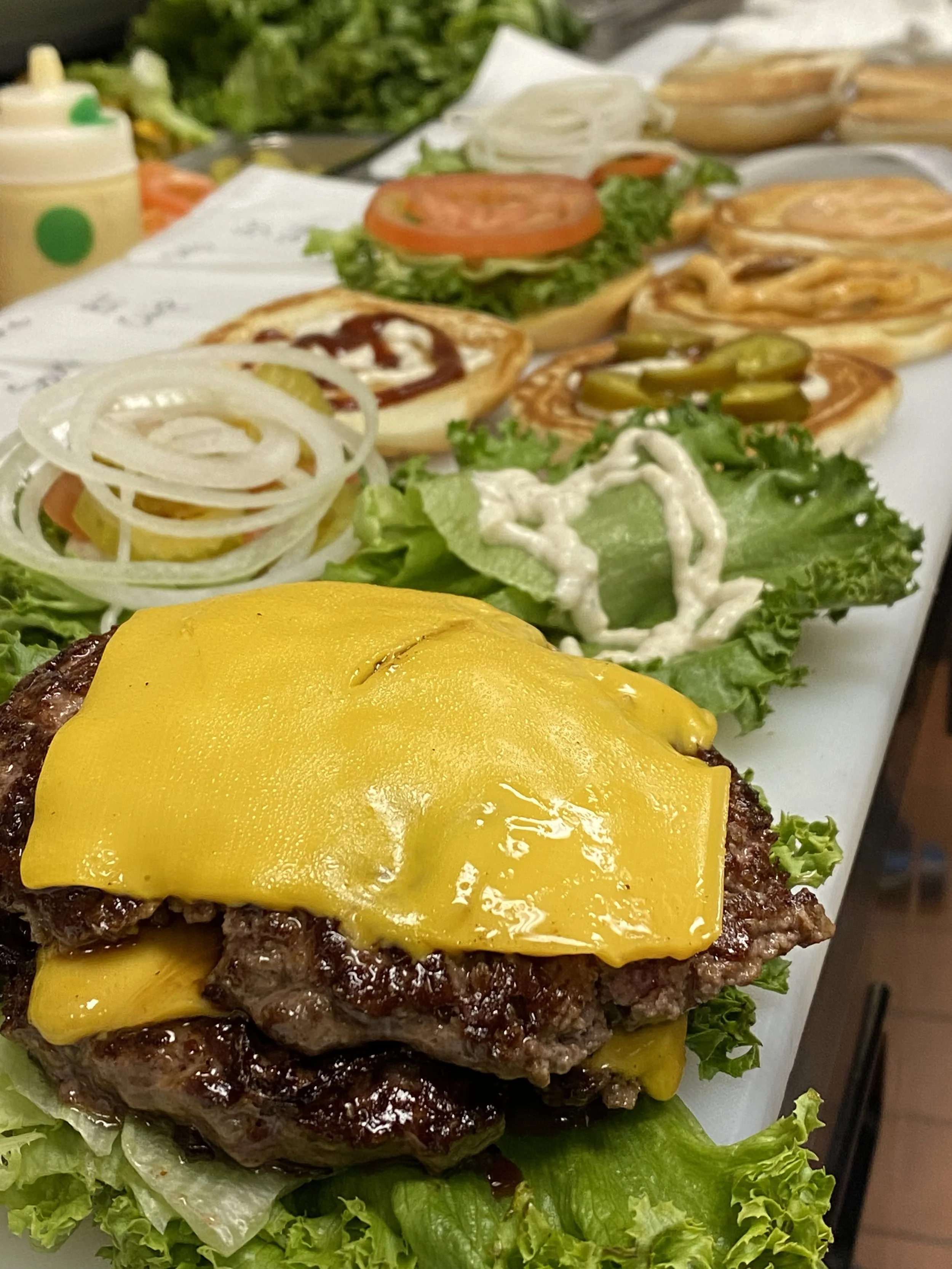 Close-up of a cheeseburger with lettuce and cheese in the foreground, with a row of mini cheeseburgers topped with different condiments and vegetables on a white surface in the background.