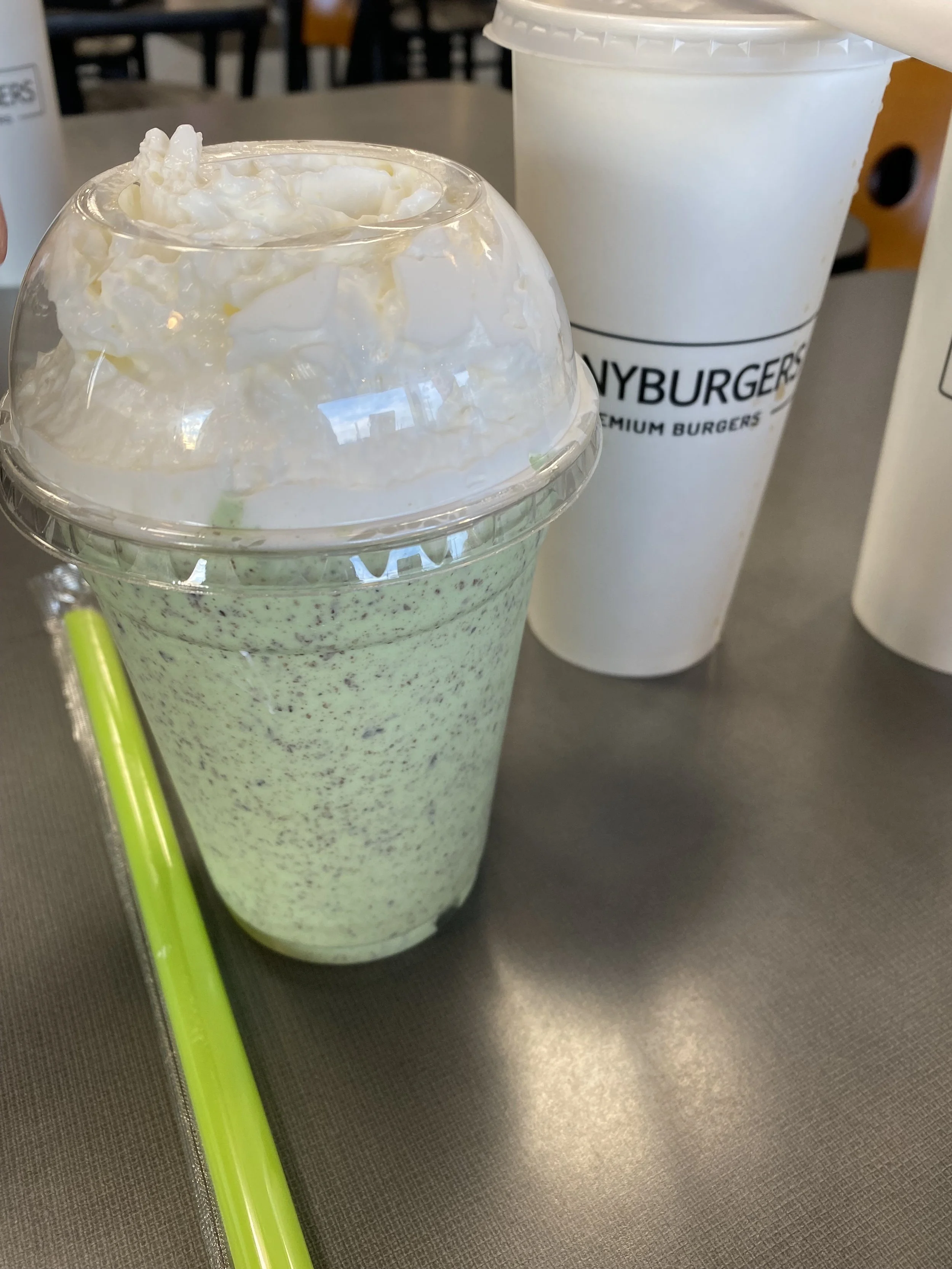 A green cookies and cream milkshake topped with whipped cream in a clear plastic cup with a domed lid, placed on a gray table next to a white cup with a black straw and a green straw