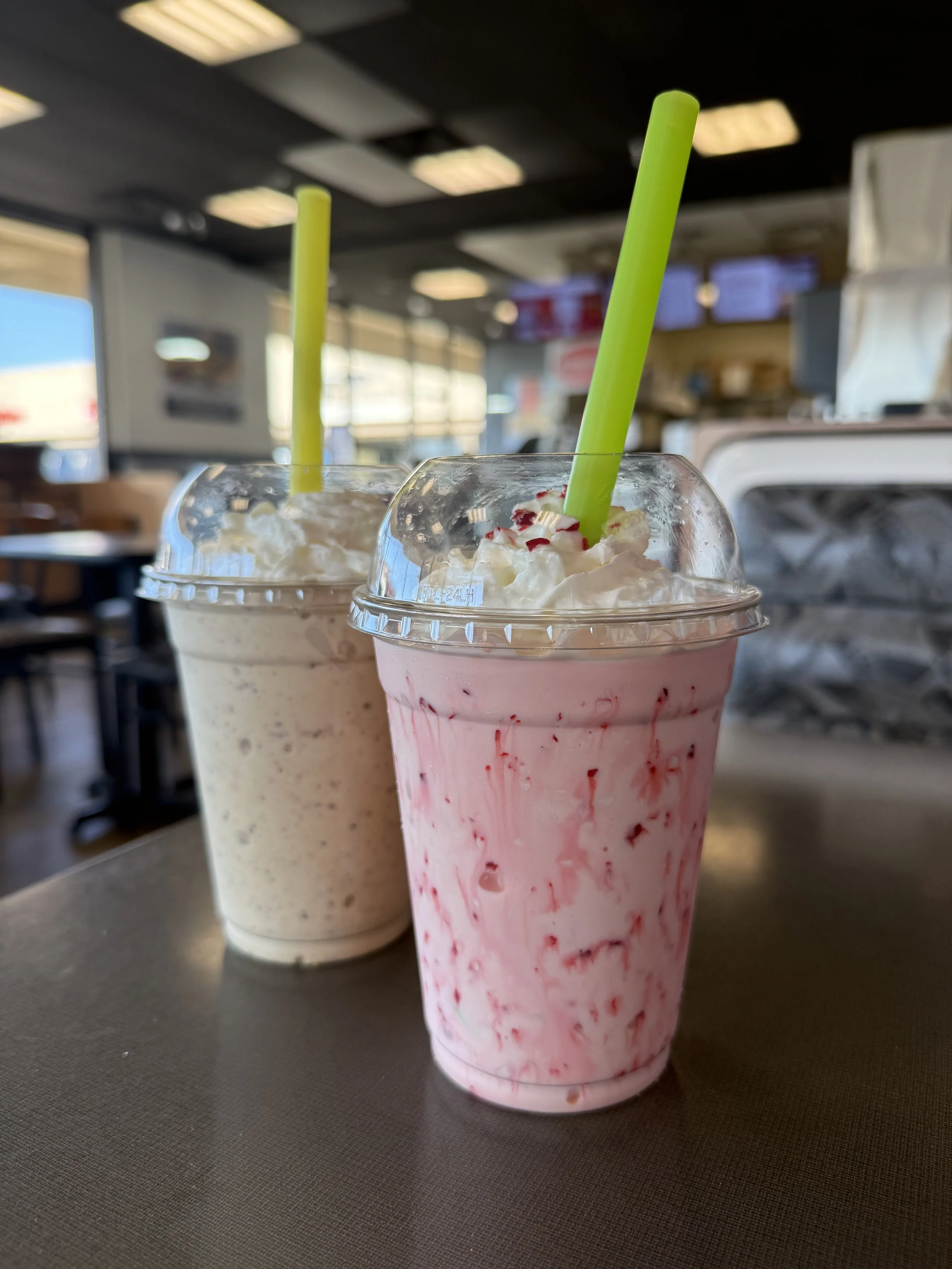 Two milkshakes with whipped cream and colorful sprinkles, one vanilla and one strawberry, in a cafe or diner setting.