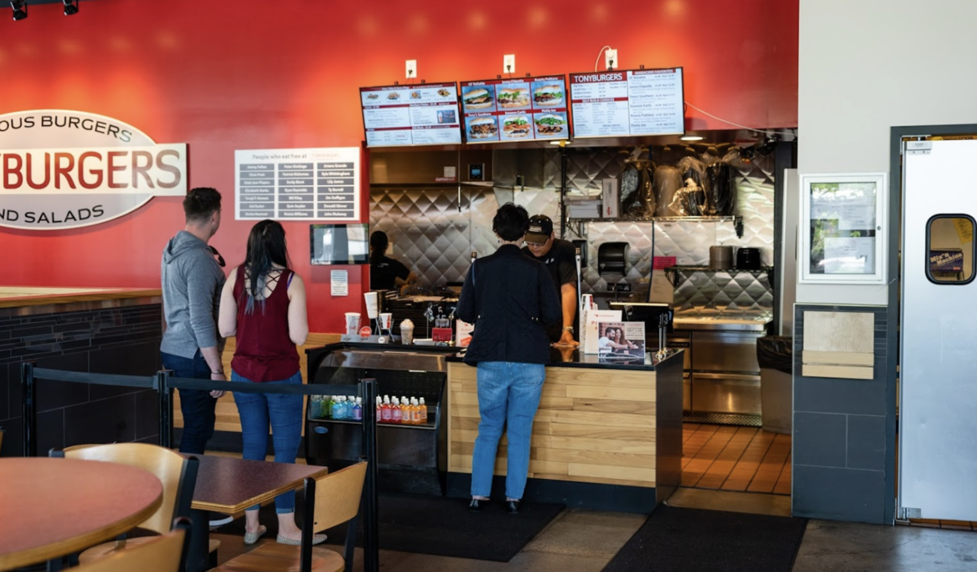 Customers ordering food at the counter of a fast-food restaurant with digital menu screens above and a red wall background.