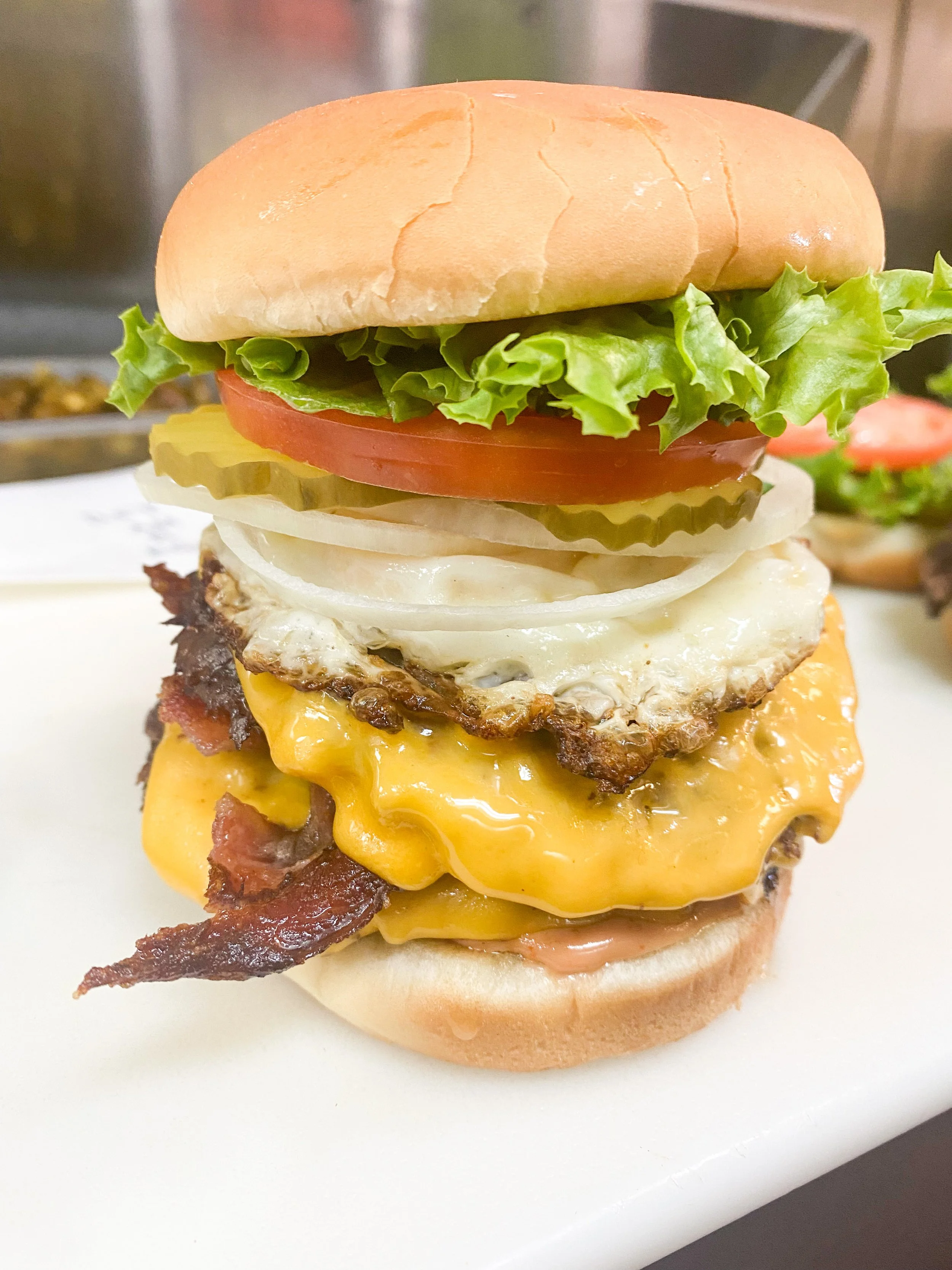 Close-up of a cheeseburger with lettuce, tomato, pickles, onions, egg, bacon, and melted cheddar cheese on a bun.