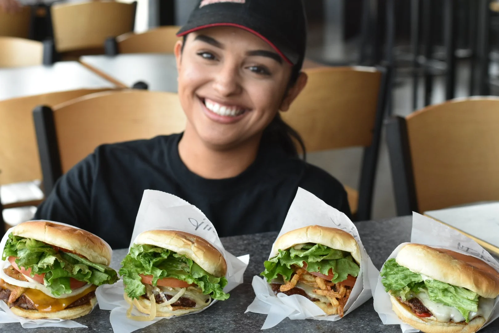A smiling woman at a fast-food restaurant with four different burgers on the counter in front of her.