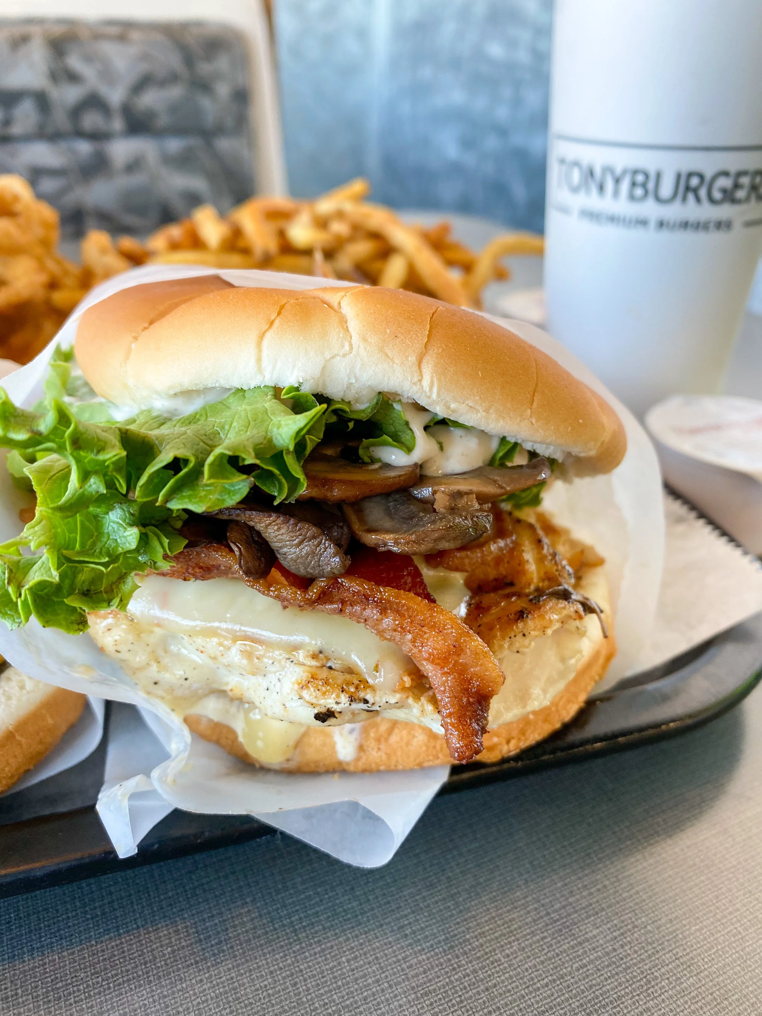 Close-up of a burger with lettuce, bacon, fried egg, mushrooms, and sauce, served with fries and a drink in the background.