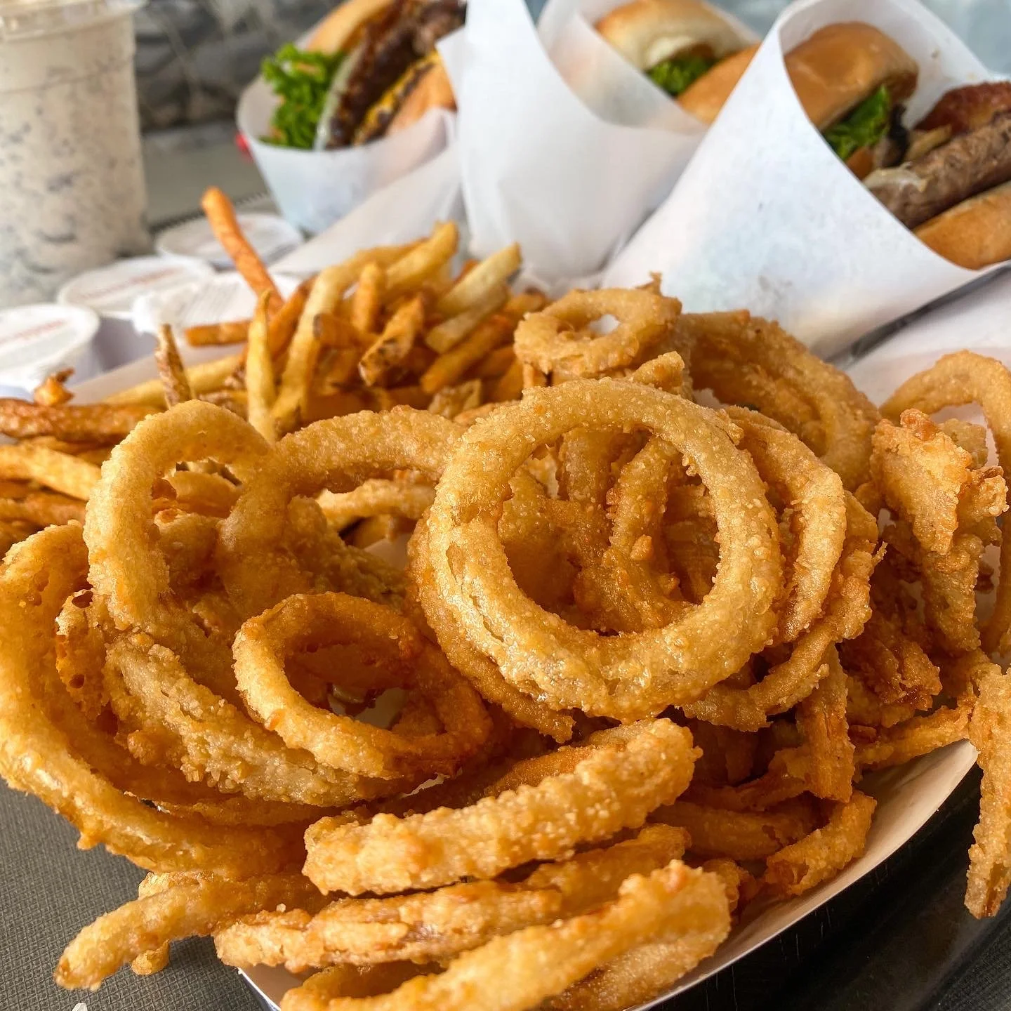 A basket of crispy fried onion rings and French fries, with cheeseburgers wrapped in paper in the background.