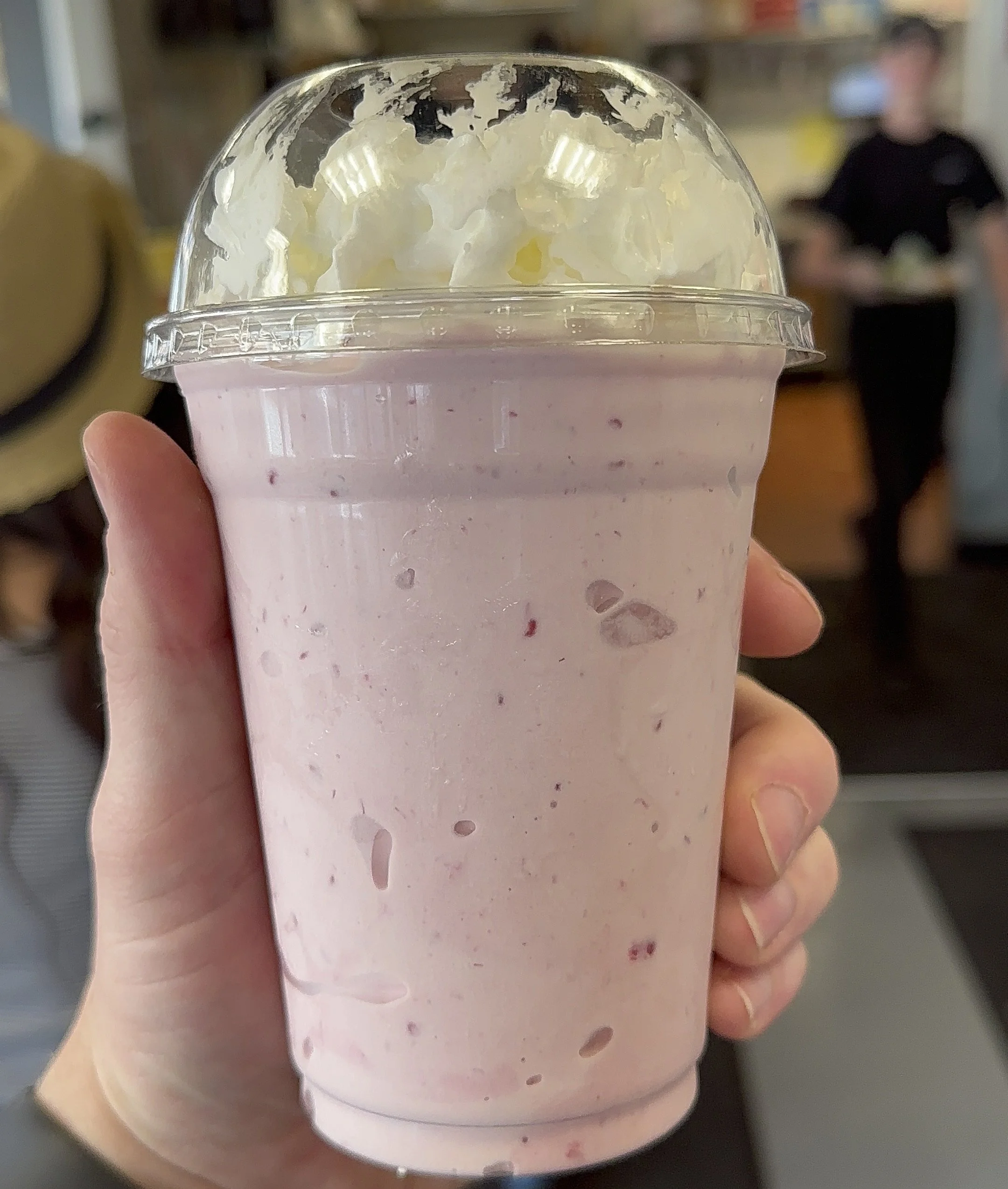 A hand holding a large plastic cup filled with a pink berry smoothie topped with whipped cream, inside a cafe